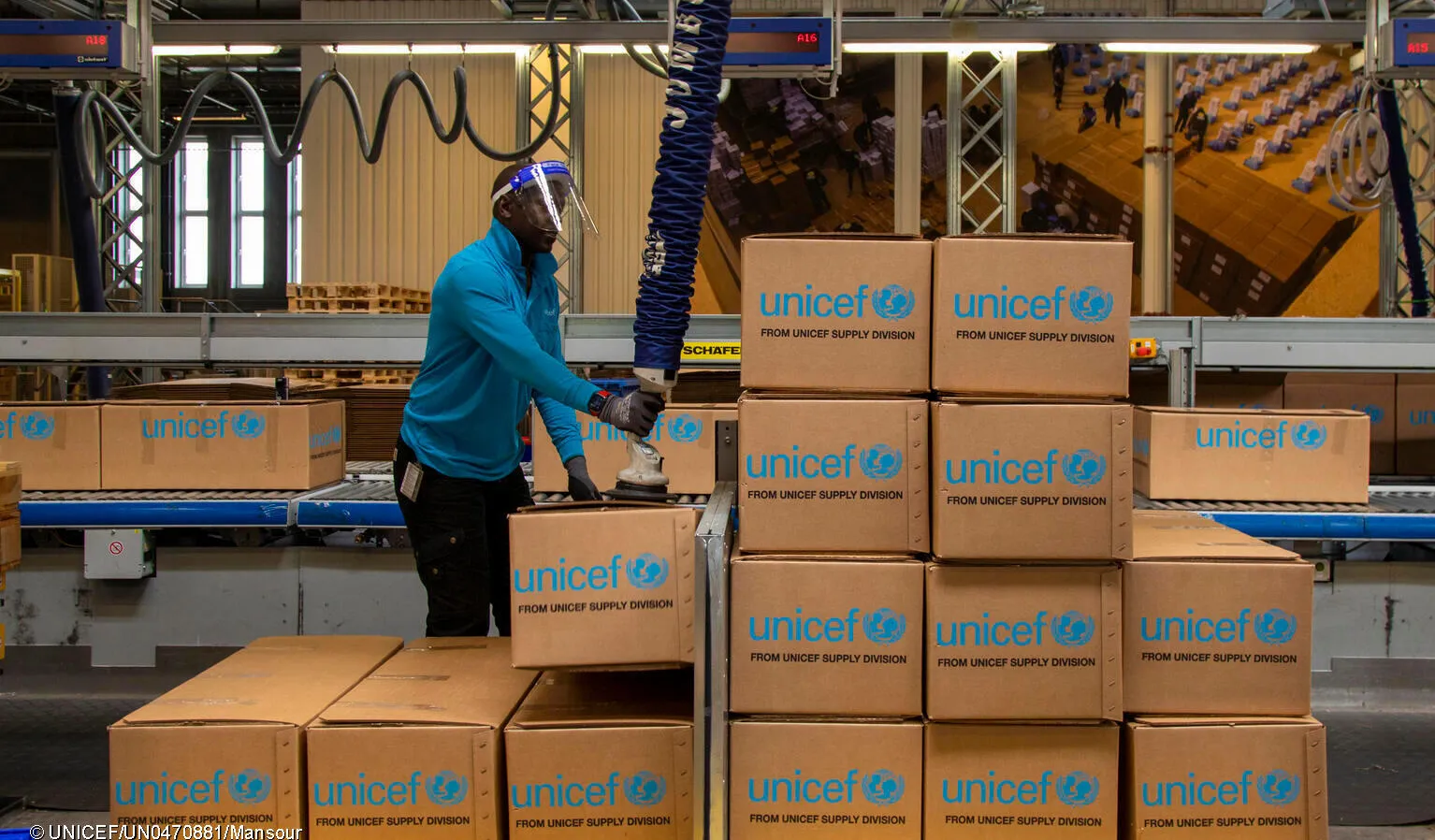 A man uses a robotic arm in a warehouse. There are boxes with UNICEF labels stacked in front of him. This photo was taken in Denmark in 2021. 