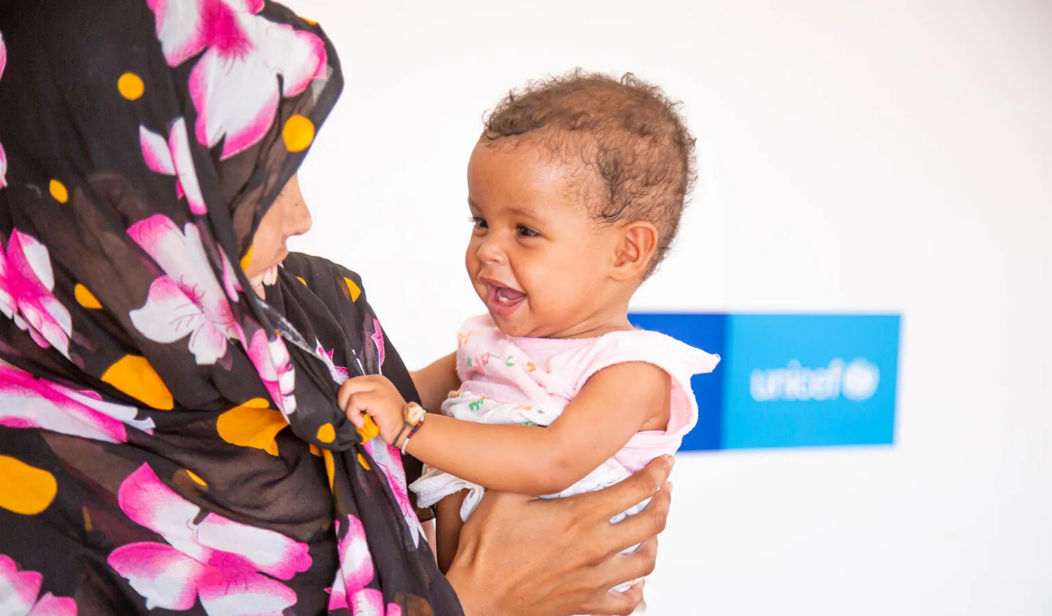 A mother holds her daughter, the mother is wearing a floral-patterned scarf and the child is wearing a pink dress. The baby and mother are both smiling. This photo was taken in Mauritania in 2021.