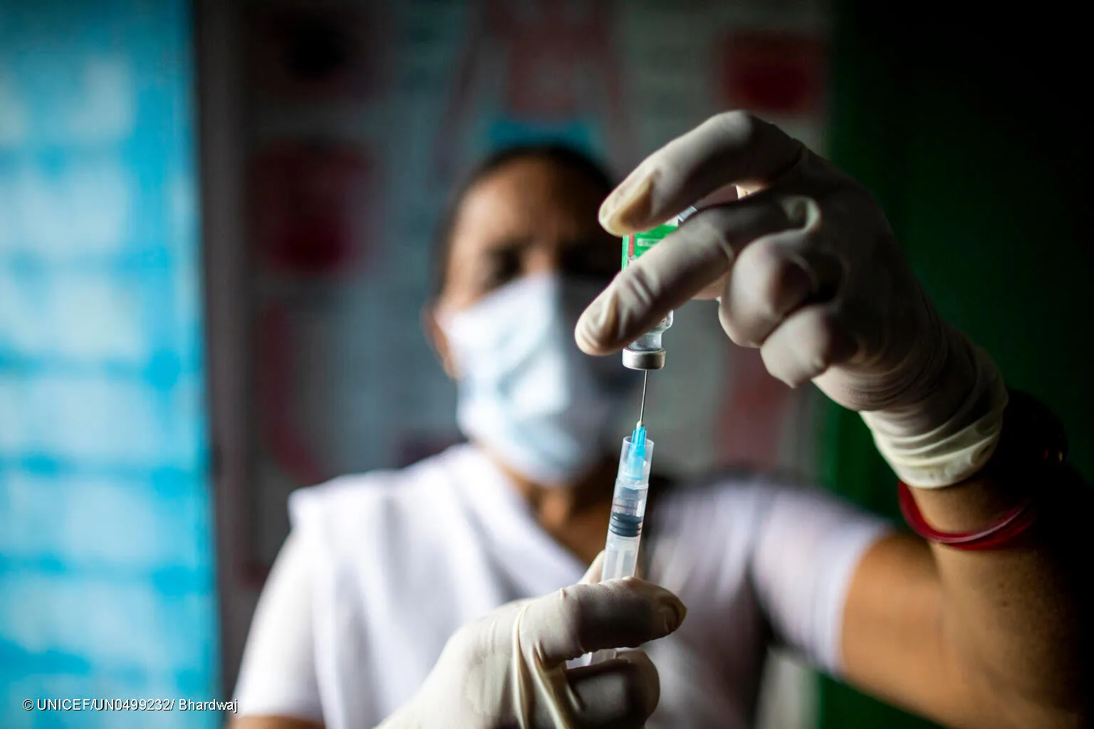 A health worker uses a syringe to extract a vaccine from its vial. He is wearing gloves. The syringe and vaccine vial and his hands are in focus and he is in the background out of focus. This photo was taken in India in 2021. 