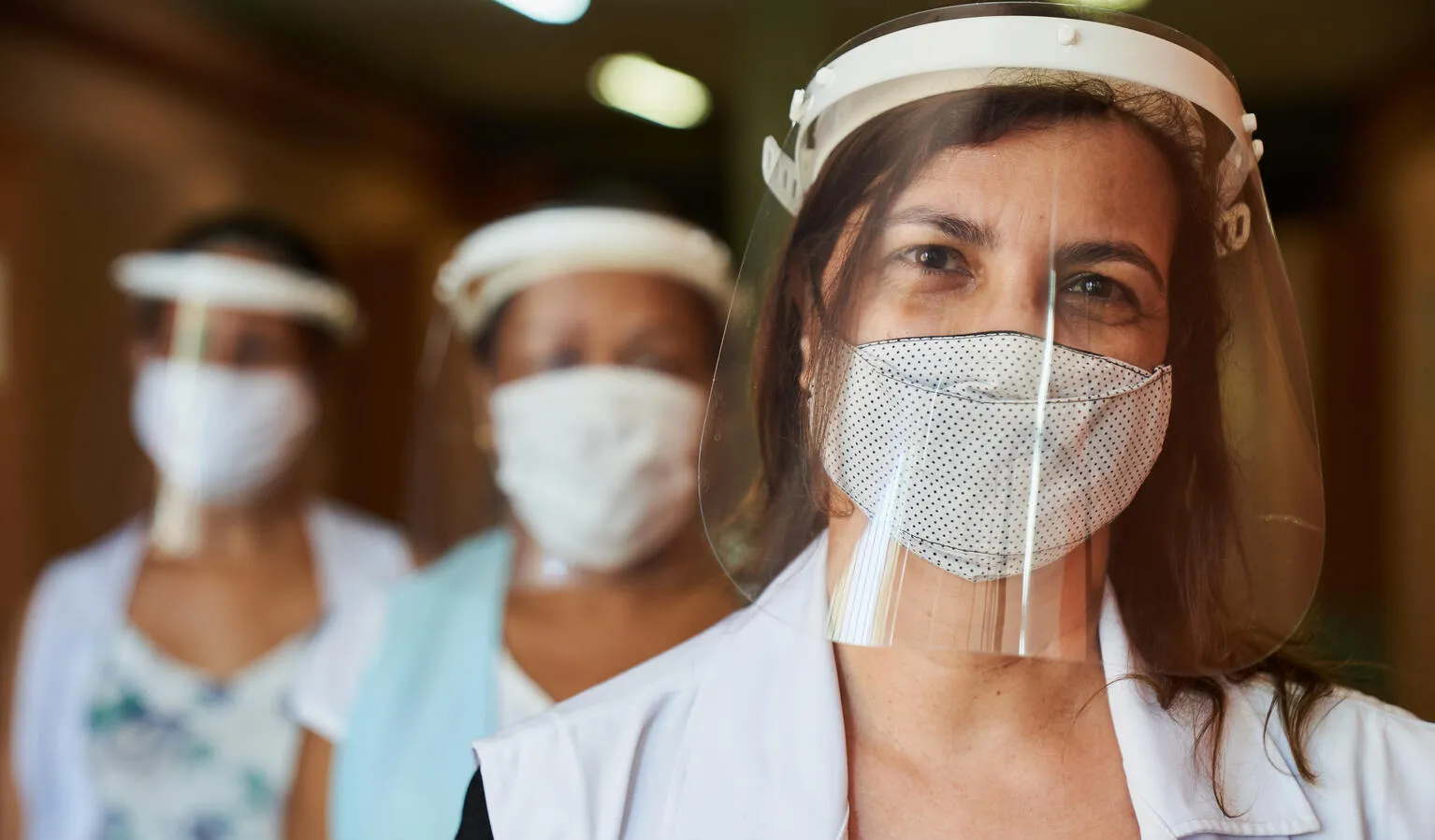 Three women stand in a line, the first one is in focus. They are all wearing masks and PPE headgear. This photo was taken in Brazil in 2021. 