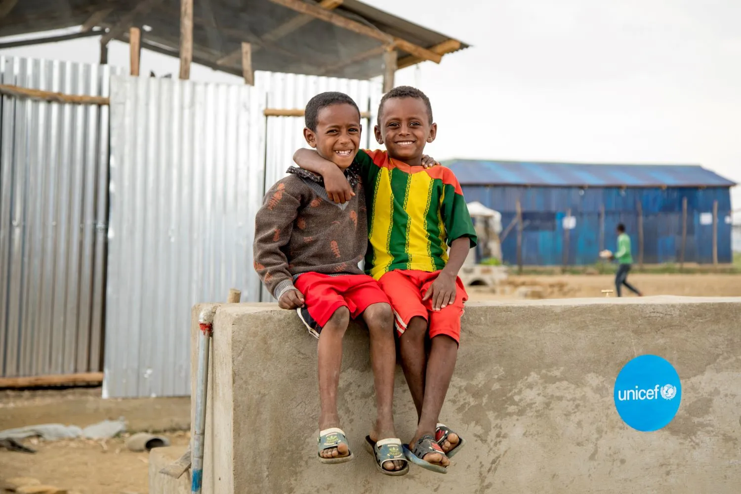 Two children sit on a concrete wall with their arms around each other’s shoulders and smiling at the camera.