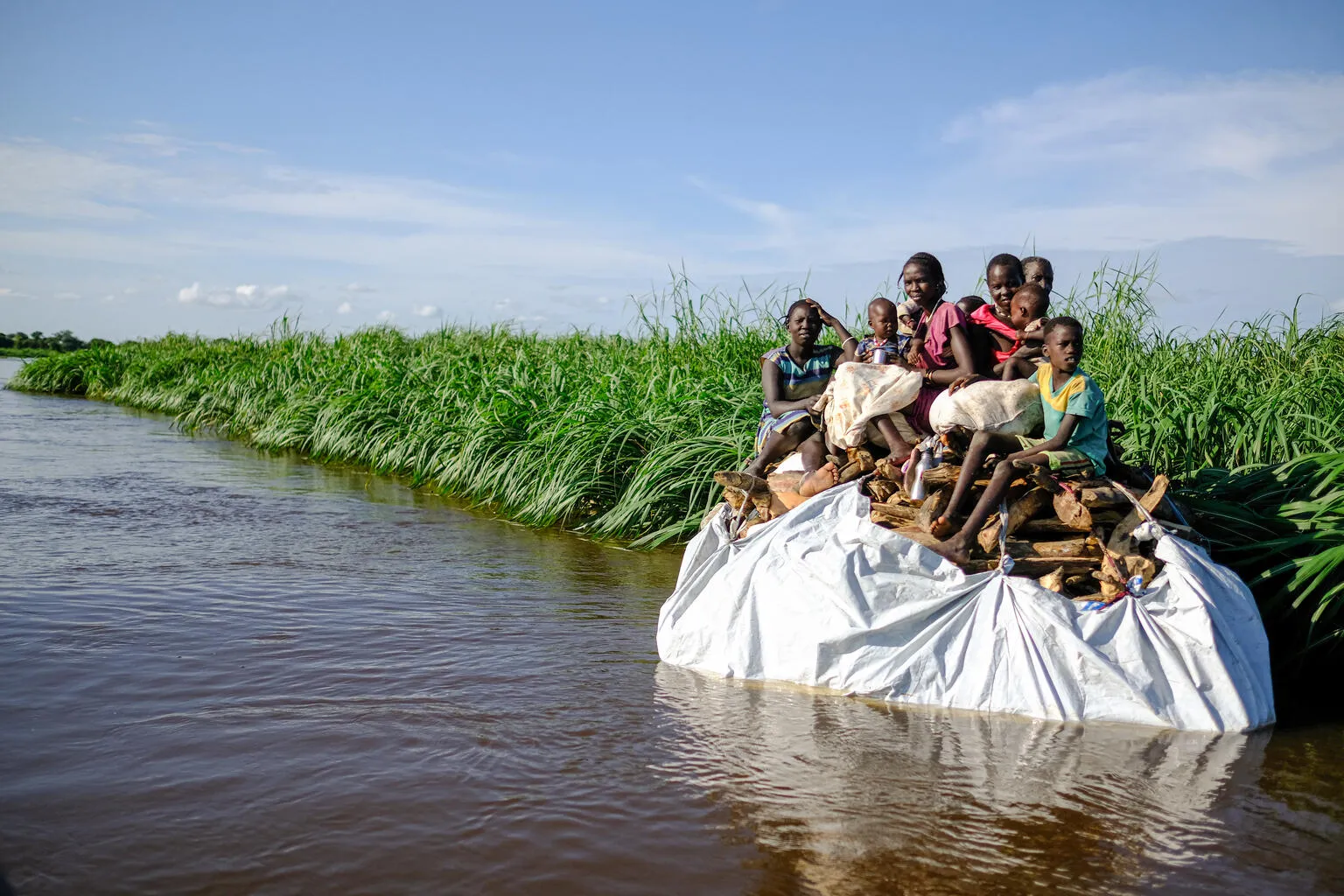 A group of children sit on top of a tarp of belongings on the sides of a flooded river bank.