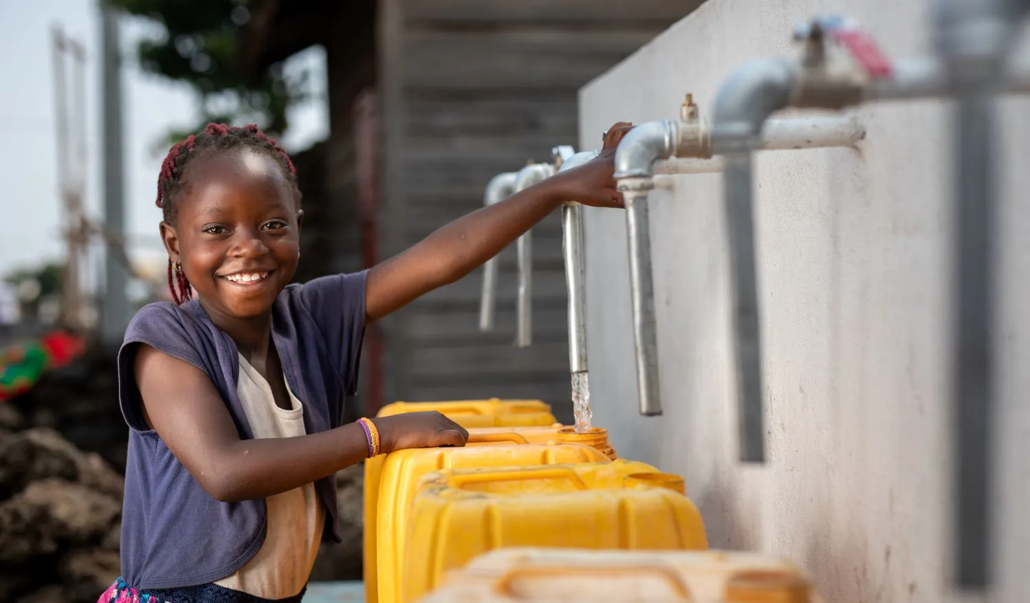 A girl smiles at the camera while filling a yellow water jug at an outdoor tap.