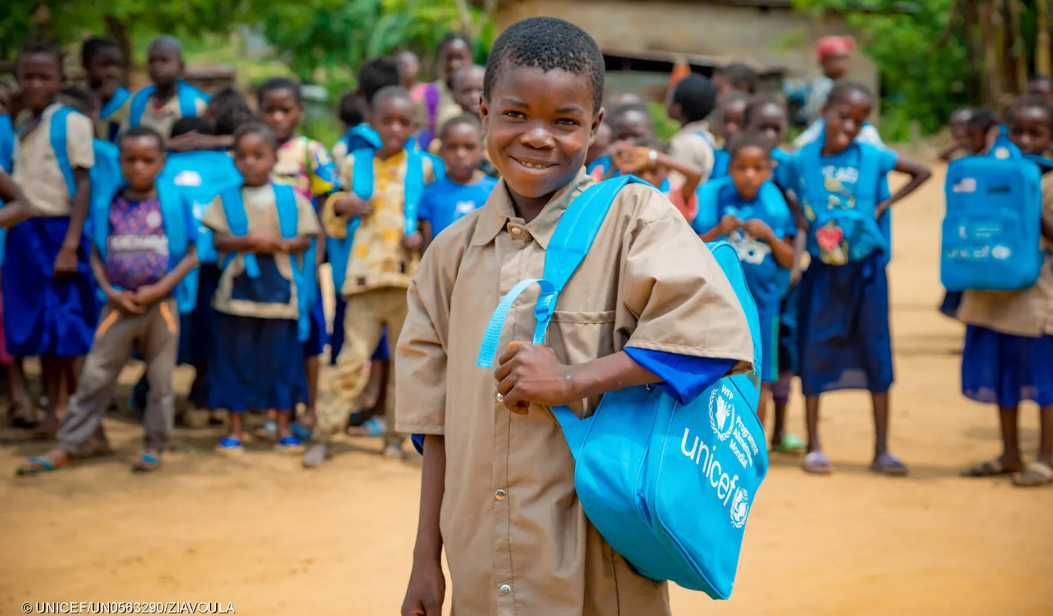 A child with a blue UNICEF bag pack slung on one side smiles at the camera. He is wearing a brown uniform, behind him out of focus are many children also wearing bag packs and brown uniforms.  This photo was taken in the Congo in the province of Lékoumou. .  