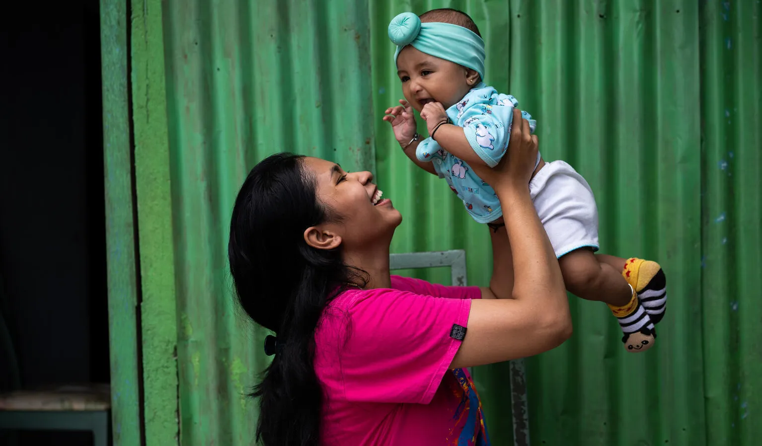 A woman wearing a pink tshirt holds her infant above her head, smiling. The infant is also smiling. 