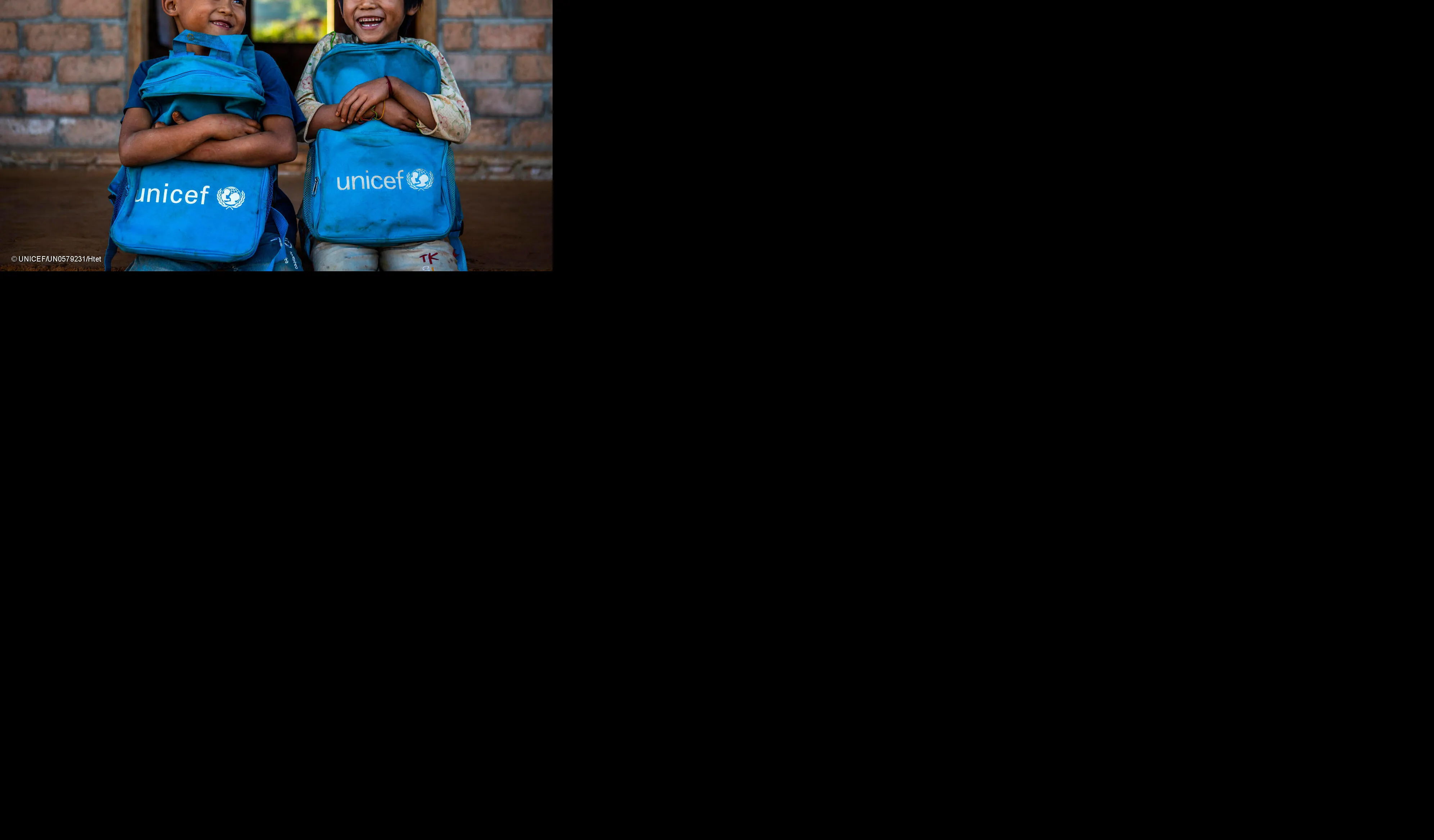 Two children holding UNICEF backpacks sit at the stoop of a classroom, smiling. 