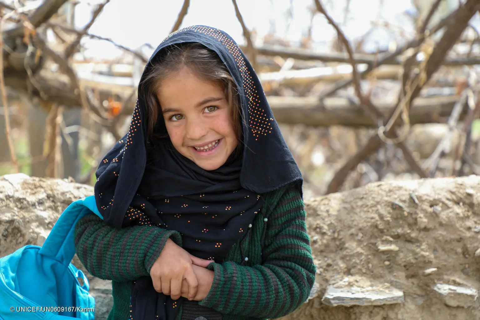 En Afghanistan, une jeune fille sourit à la caméra, la sangle bleue d’un sac à dos de l’UNICEF sur le bras.