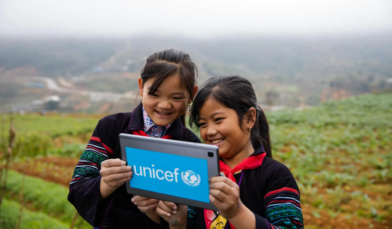 Two girls stand outdoors in a mountainous green field, looking at a tablet with a UNICEF logo on it. This photo was taken in Vietnam. 