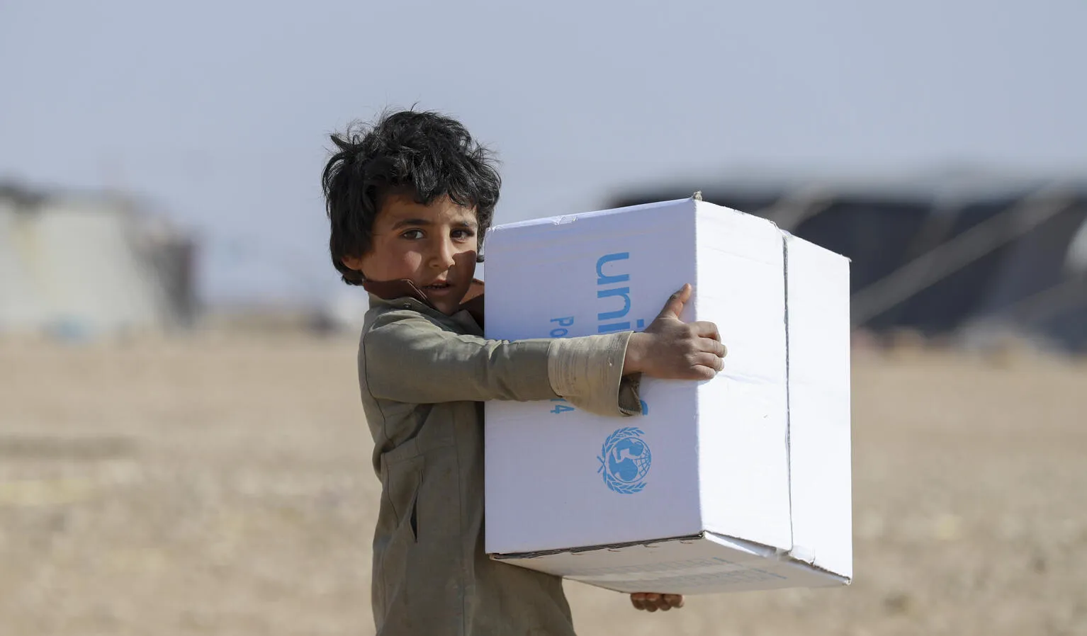 A child holds a UNICEF box, in the background out of focus you can see tents