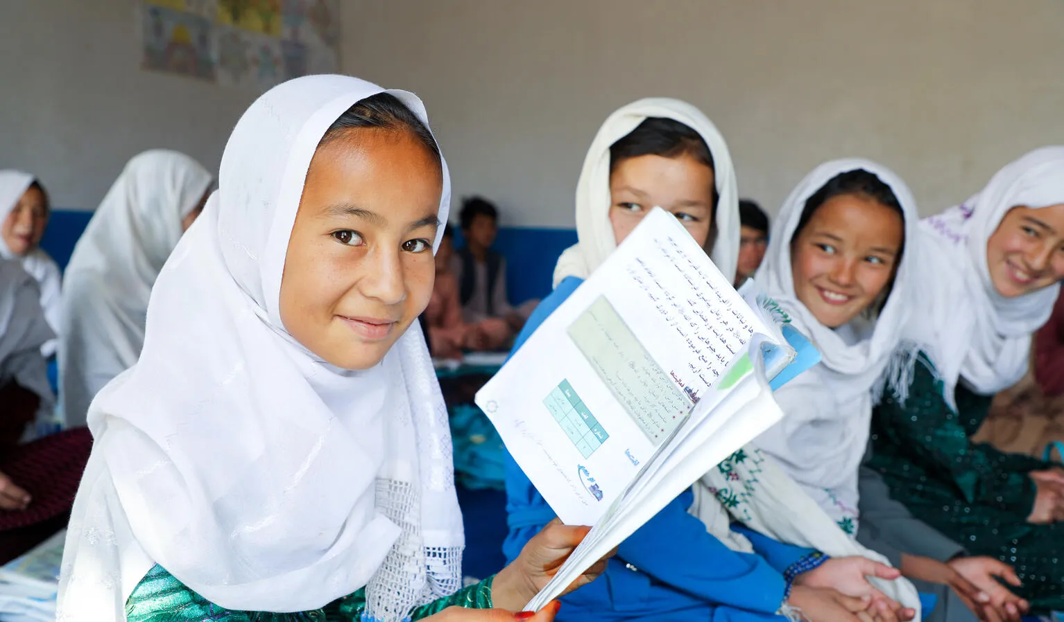 At a classroom in Afghanistan, a girl wearing a white headscarf holds open a book and smiles at the camera.