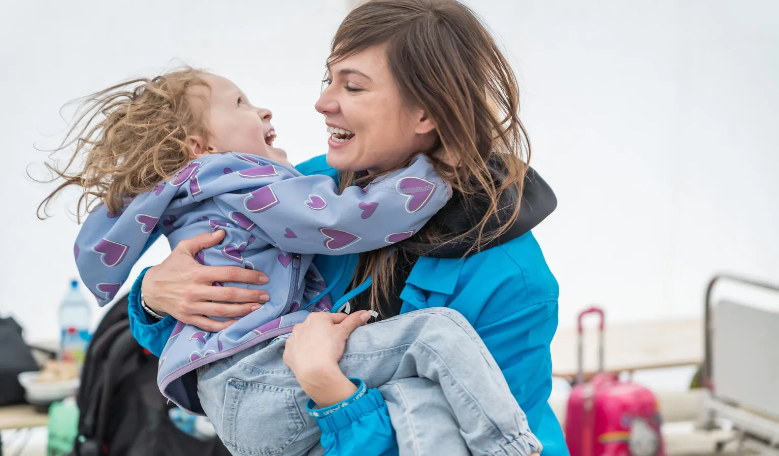 A woman wearing a UNICEF jakcet carries a child in her arms, both the woman and child are laughing. They are outdoors and both are wearing jackets.