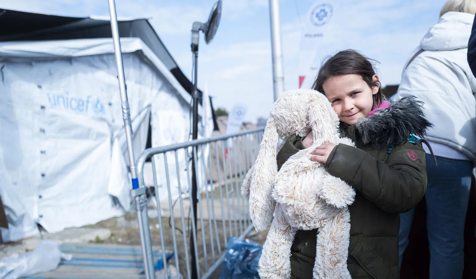 A child holds a bunny as she looks at the camera with a small smile. She is wearing a jacket and standing outdoors, in a line, behind her are white UNICEF tents. This photo was taken at the Ukrainian border in 2022. 