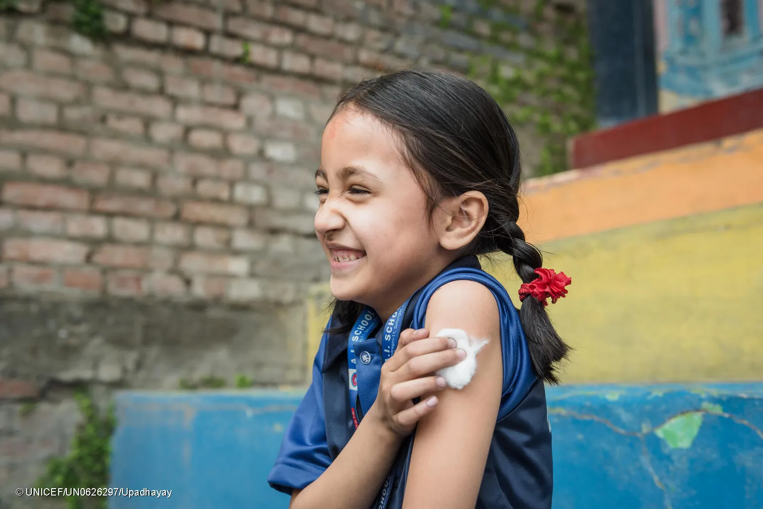 A girl holds a piece of cotton up to her recently vaccinated arm. She is smiling.  This photo was taken in Nepal in 2022. 