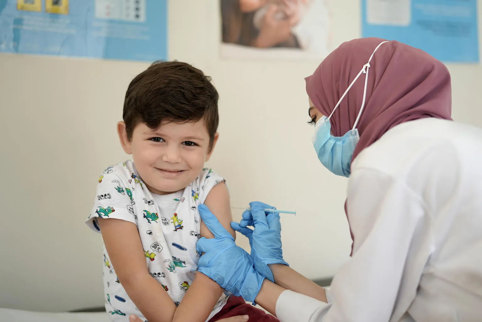 A child looks at the camera with a shy smile, a health care worker in a mask and medical gloves gives him a shot in the arm. The setting looks like a clinic. 