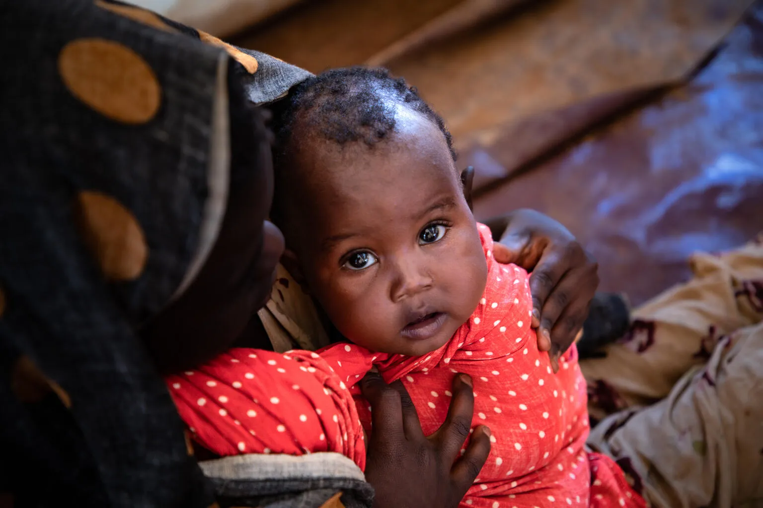 A child wrapped in a red polka dotted blanket looks up to the camera. he is held by his mother whose face is cast down.