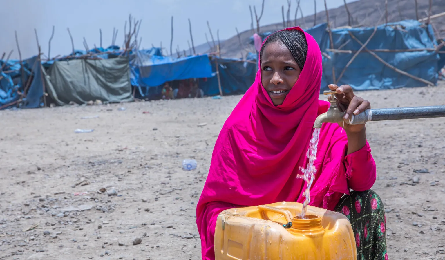 A girl in a bright pink scarf in front of yellow jerry can which has water pouring in it from a single tap. Tents can be seen in the background, looking like she is in a camp of sorts. The girl is looking away from the camera and is smiling. 
