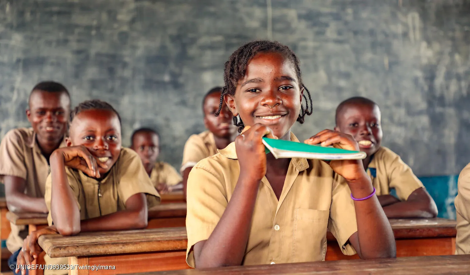 A girl sits in a classroom smiling. She is holding up a green notebook. Behind her are other children in brown uniforms, also smiling. This photo was taken in the Congo in 2022. 