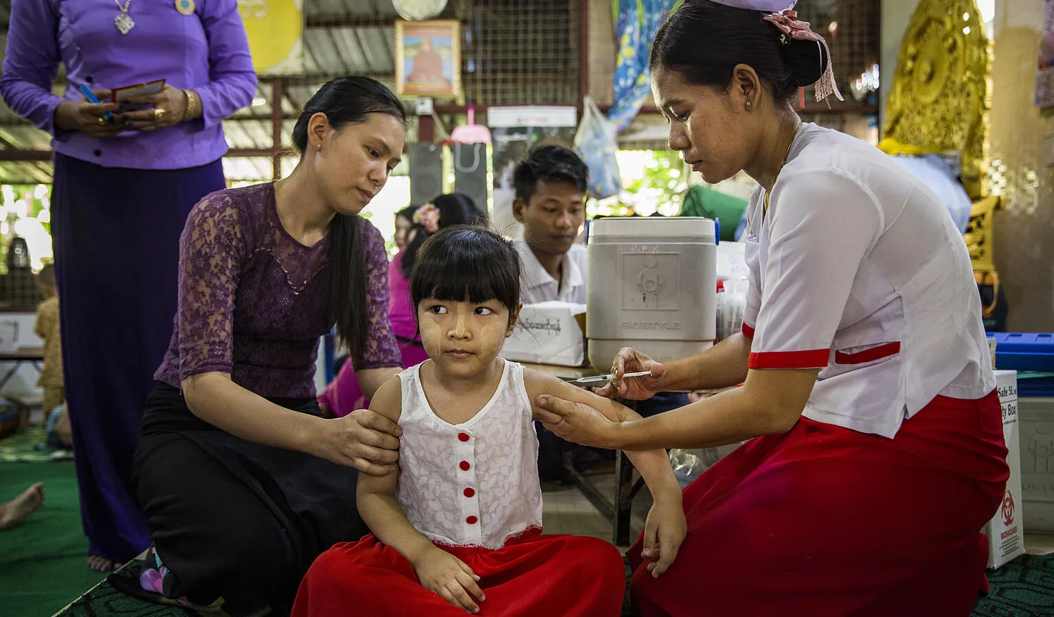 A girl sitting cross legged, looking very calm while getting an injection by a nurse, another woman is next to the child, looking at her. 