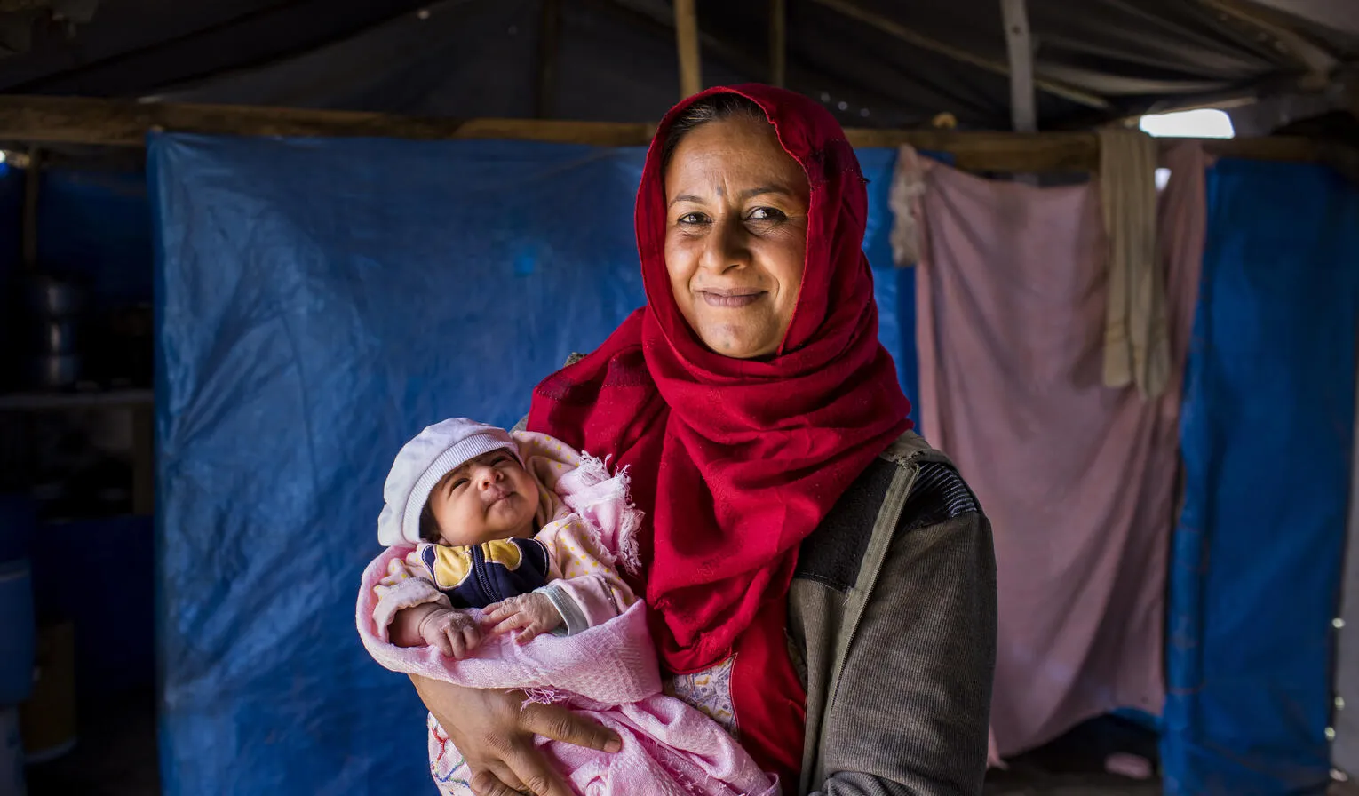 A mother in front of a tent holding a newborn in pink blanket. 