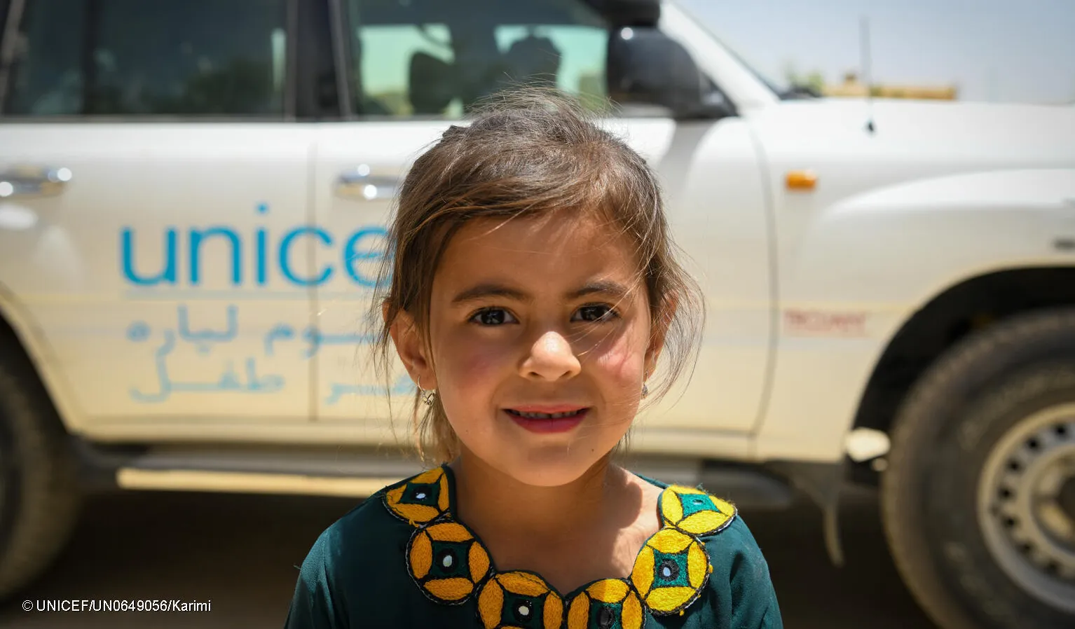An Afghan girl looks at the camera with a shy smile, behind her is a white vehicle with the words UNICEF written on it. 