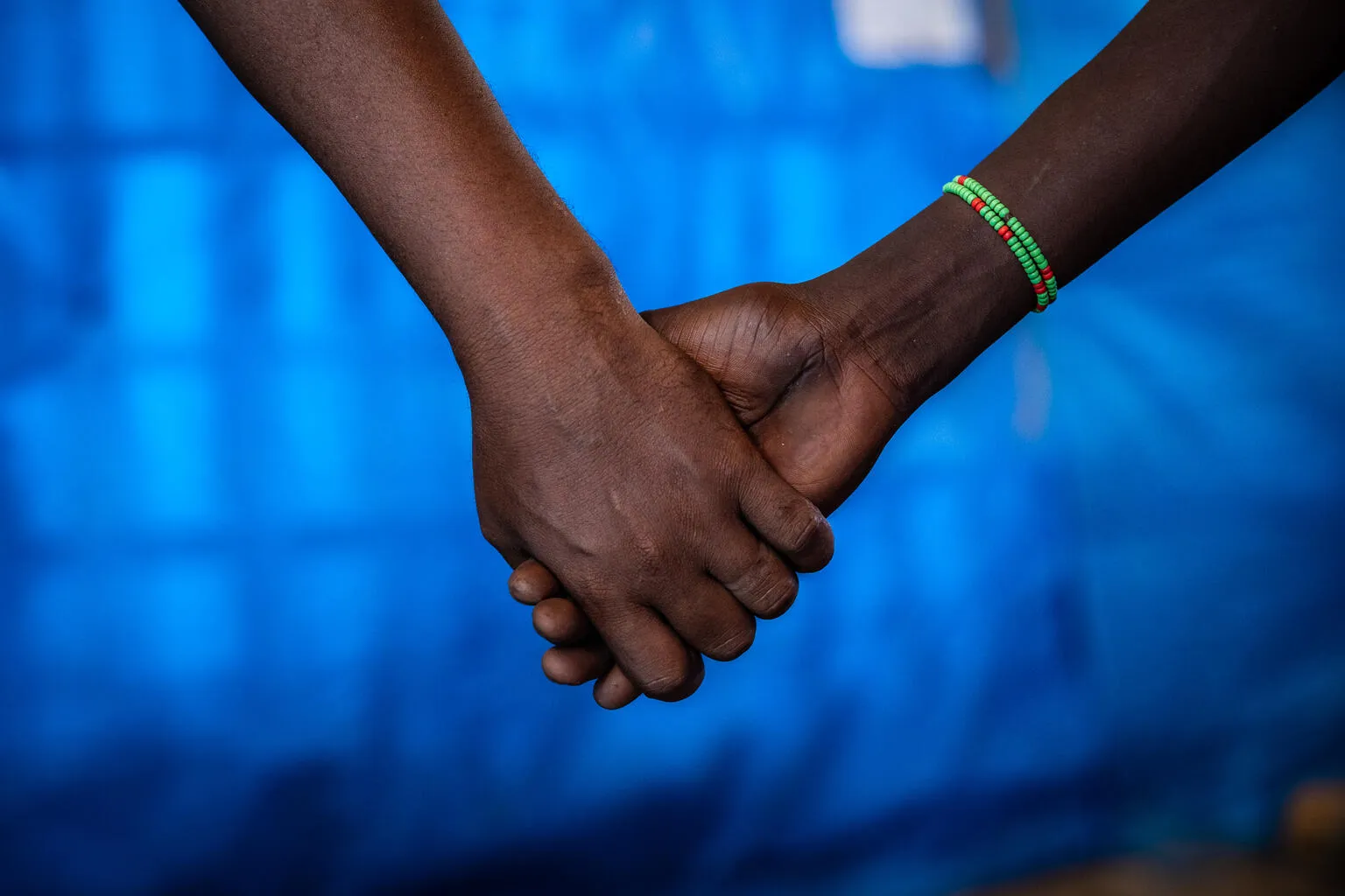 Hands being held, the hand on the left is that of a man and the one on the right of a girl. The girl is wearing a green beaded bracelet. The photo is cropped till the forearms, the background is that of a blue tarp.