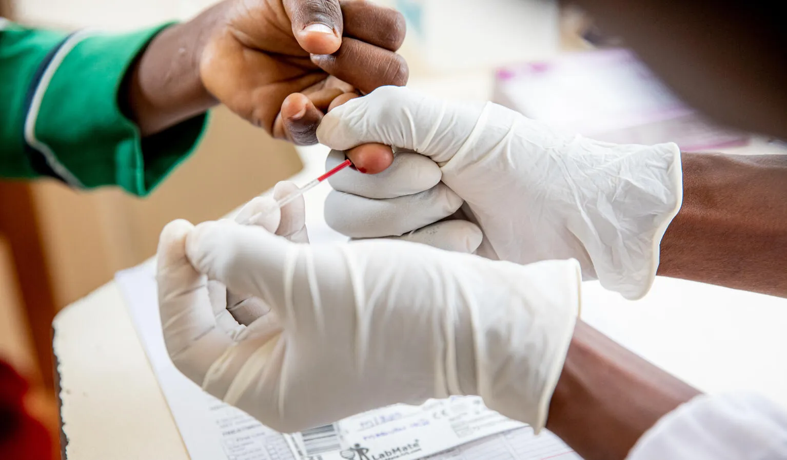 A close up of a child's hand being pricked by a medical professional's hands who is wearing white latex gloves. 