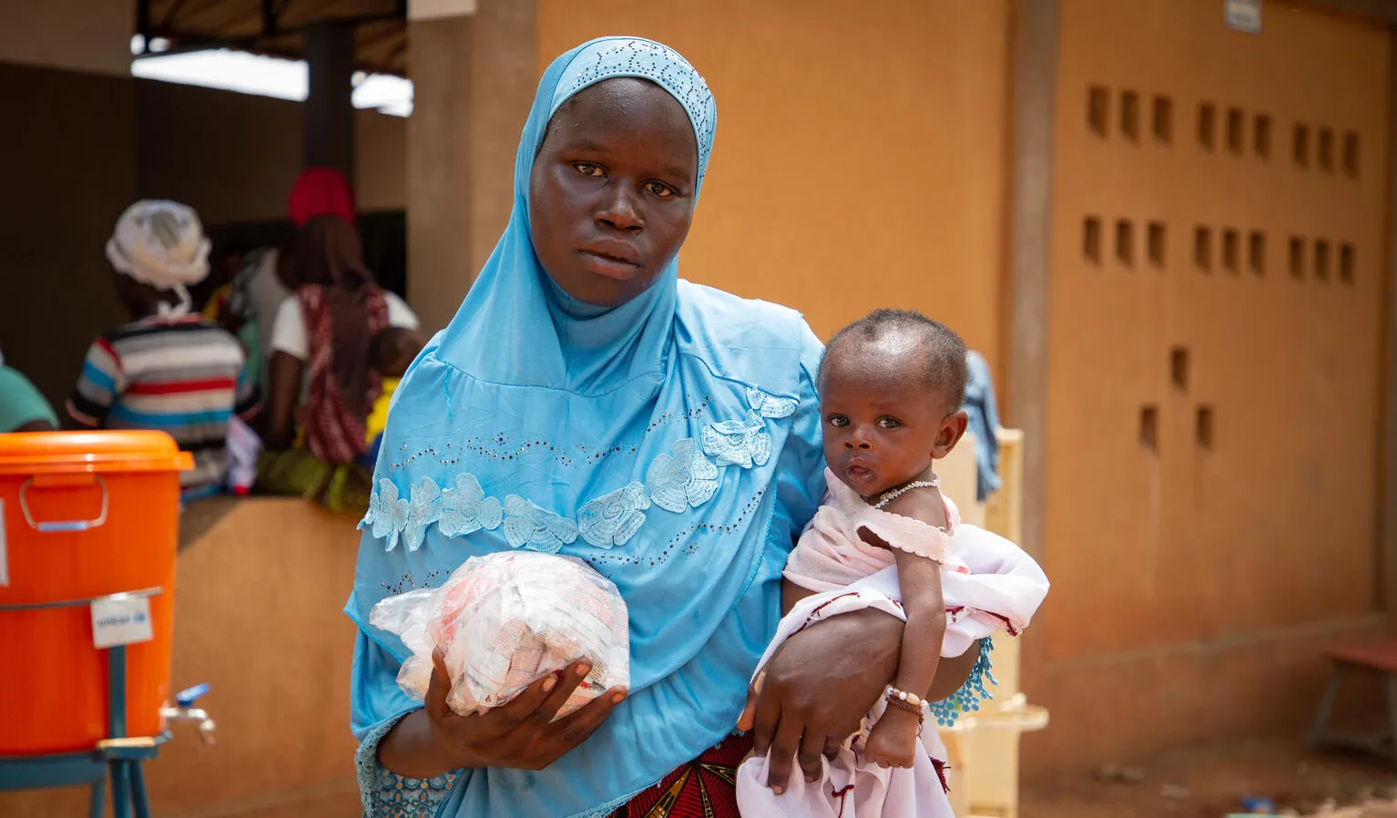 a woman in a blue scarf holding a packet of food in one hand and a child in the other. 