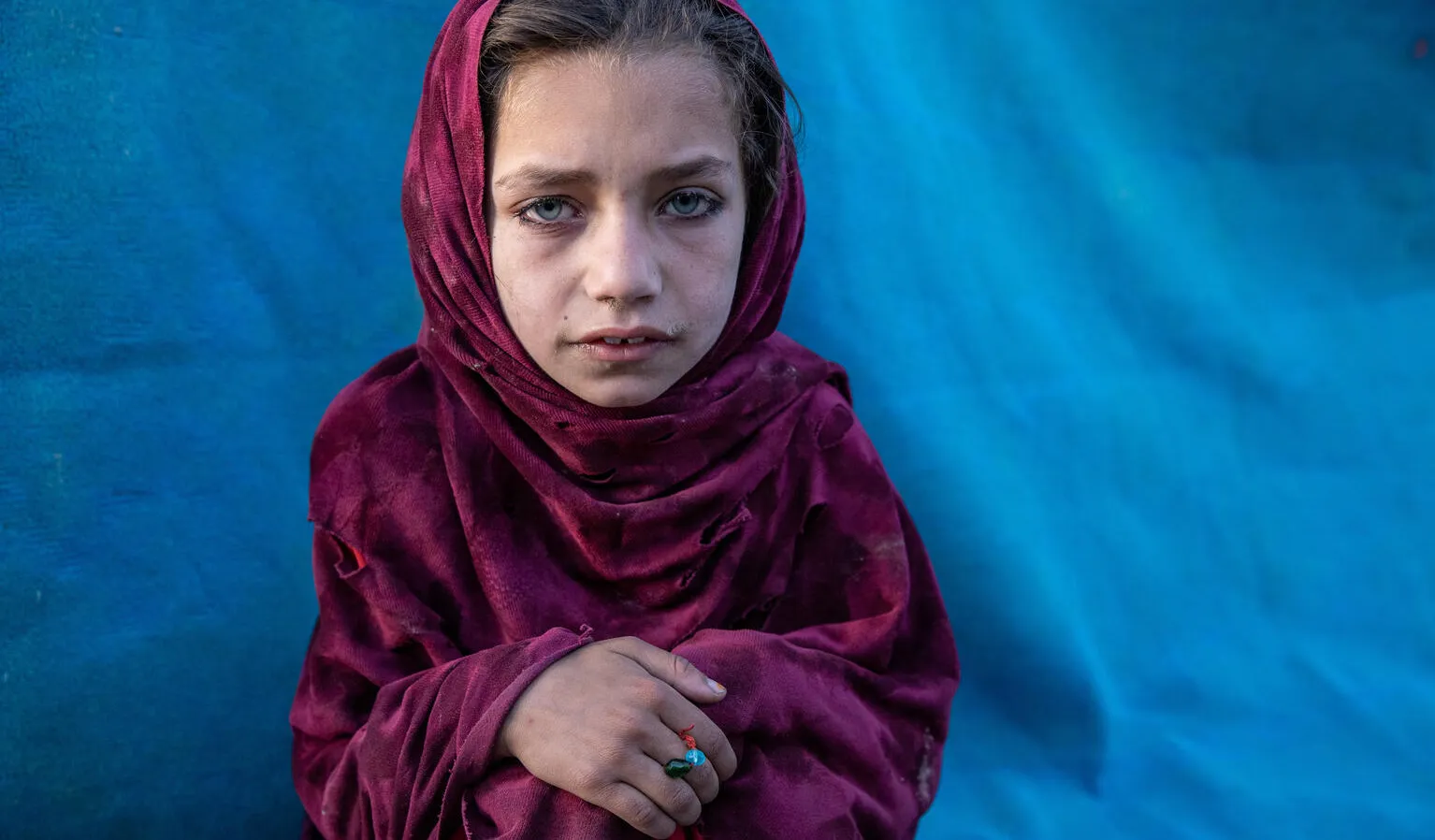 A girl wearing a magenta headscarf while sitting, looking at the camera, with her hands folded on her knees. There is a vivid blue tent behind her