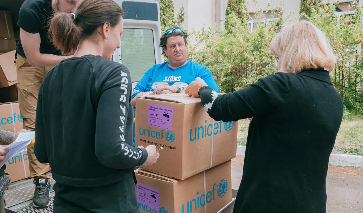 A van with the back door open and a several boxes with the words UNICEF written on them. Two women stand in front of the boxes looking at them. One man is unloading the boxes and two other men are standing to the side.