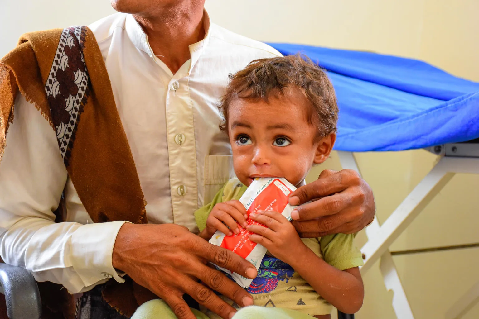A man holds a child who is eating from a red packet of Plumpy'Nut. This photo was taken in Yemen in 2022. 