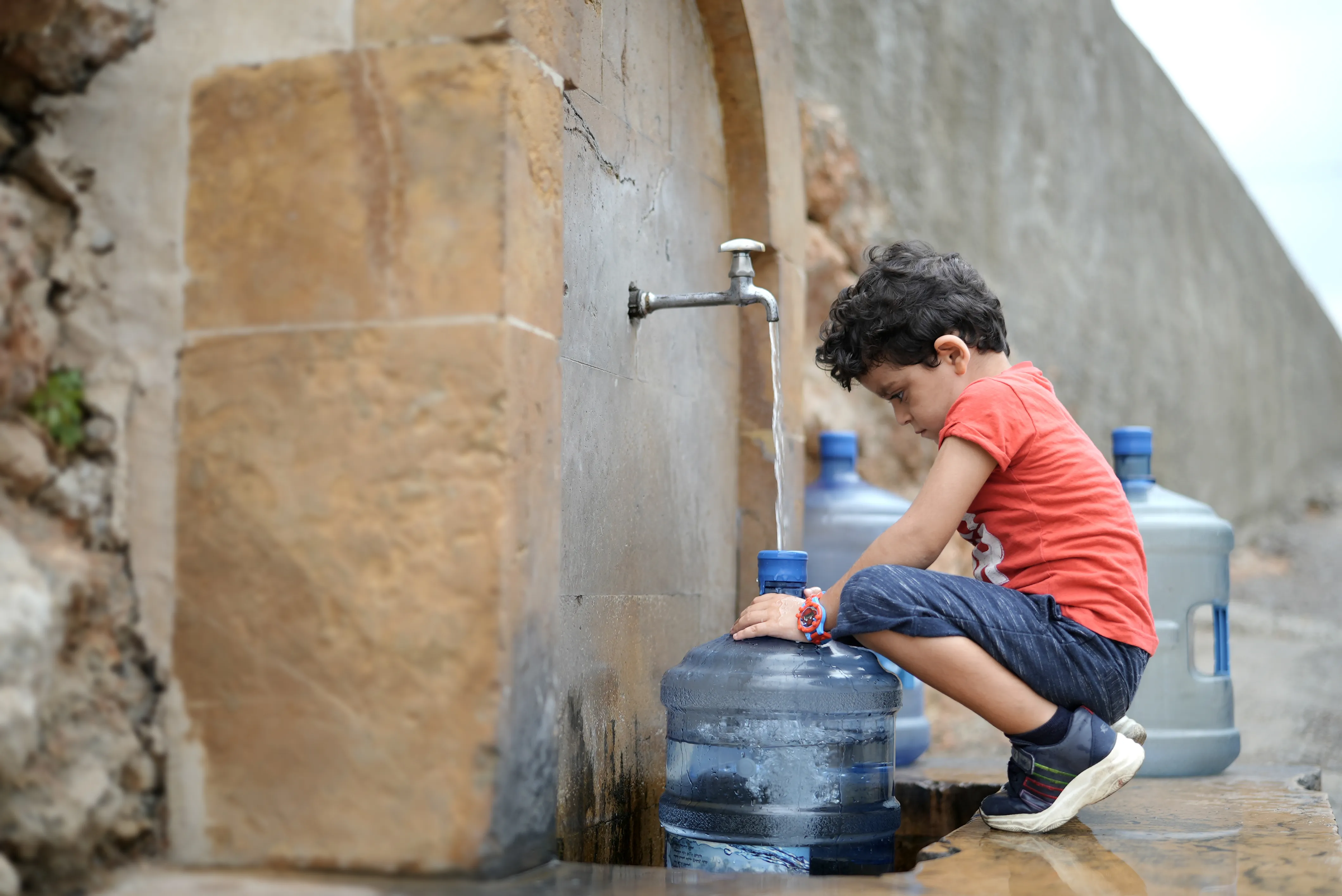 Boy crouching beside a stone fountain, filling a large water jug from a tap.
