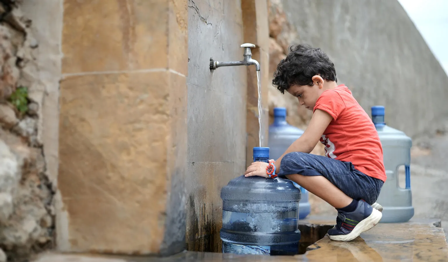 a boy squats in front of a water tap which is filling up a gallon bottle with water. 