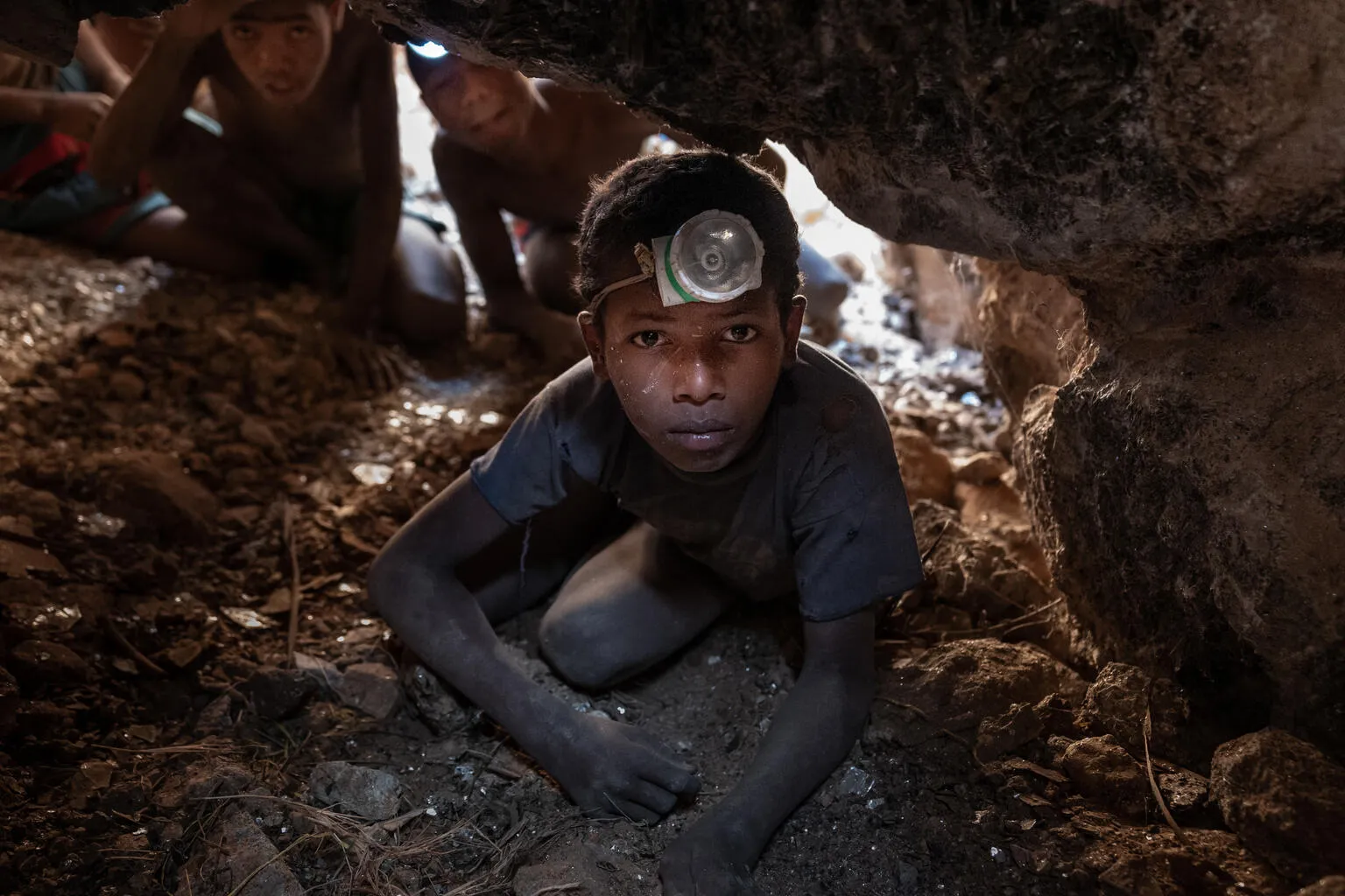 A young person wearing a headlamp crawls through a narrow, rocky cave. Behind them, others follow. The atmosphere is tense and focused.