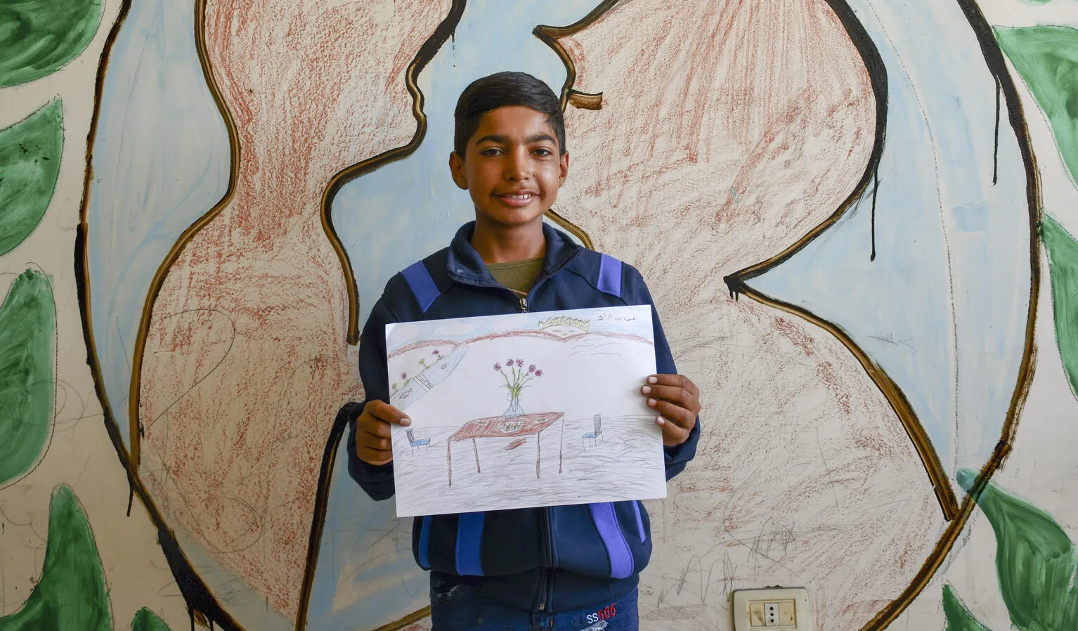 A boy holds a drawing up. It is made on paper and with colour pencils, showing a table with a vase and flowers on it. The boy is looking at the camera smiling. 