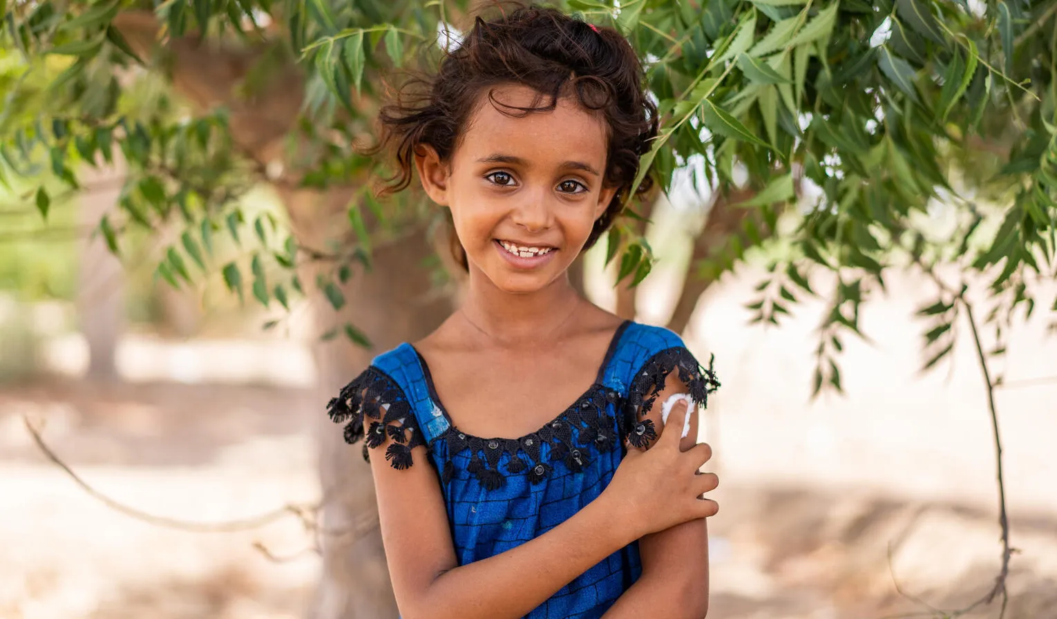 A girl in a blue dress stands under a tree smiling while holding onto a her arm where she has just been given a vaccine shot. This photo was taken in Yemen in 2022. 