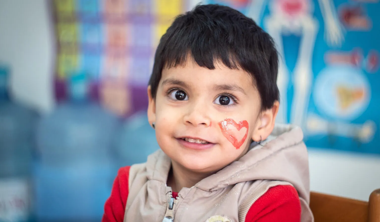 A child with a red heart painted on their face looks at the camera and smiles. They are sitting in a classroom setting. This photo was taken in Bosnia and Herzegovina in 2022.. 