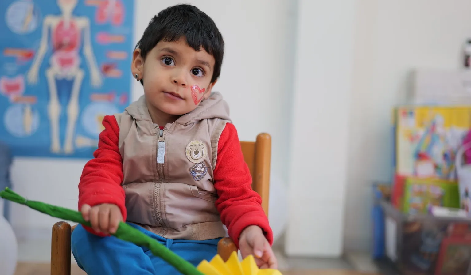 A child with paint on his face, in a classroom-like setting sits on a chair and looks directly into the camera.