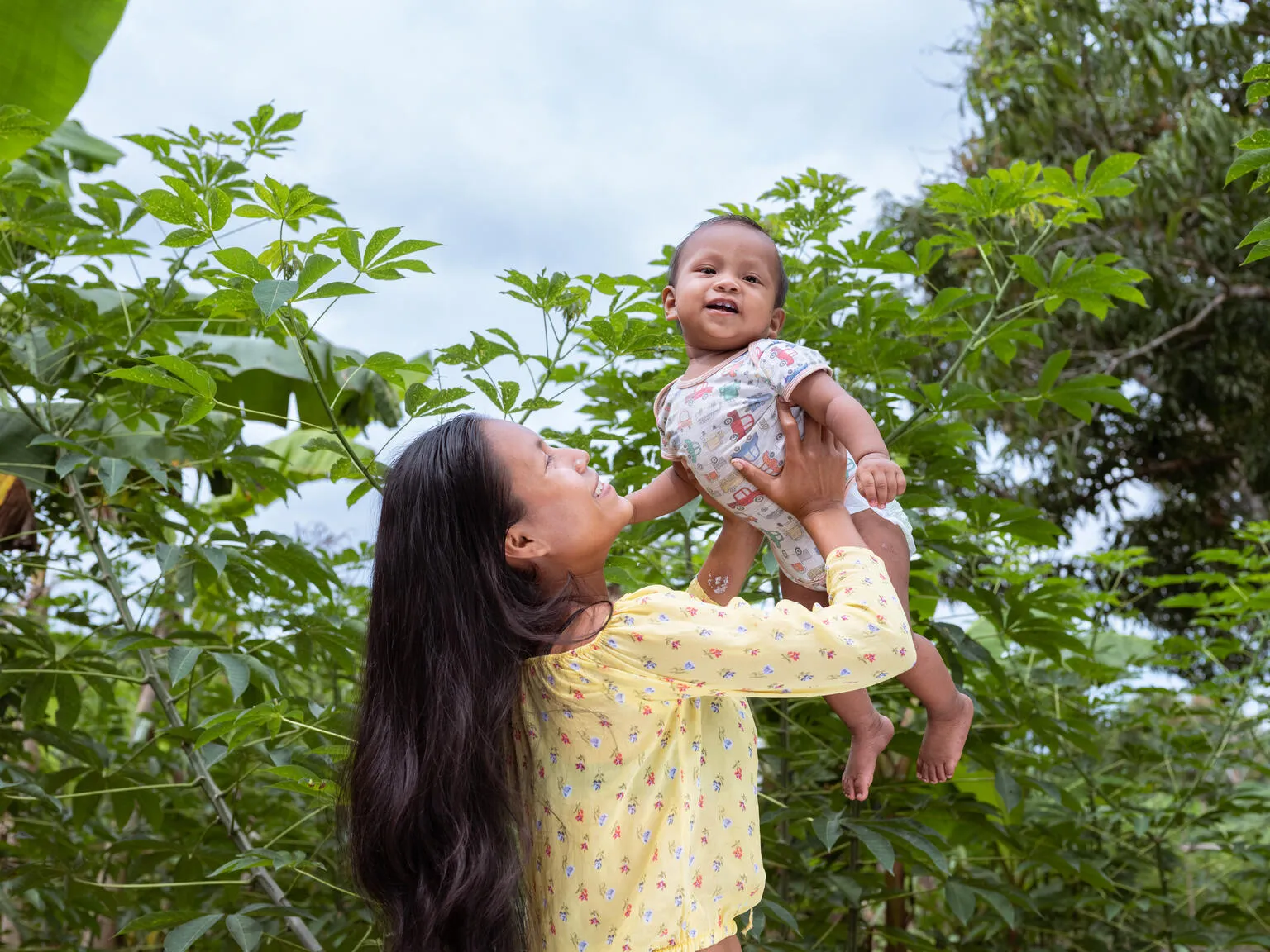 A woman hold up a baby in her arms above her head, behind her are many trees.