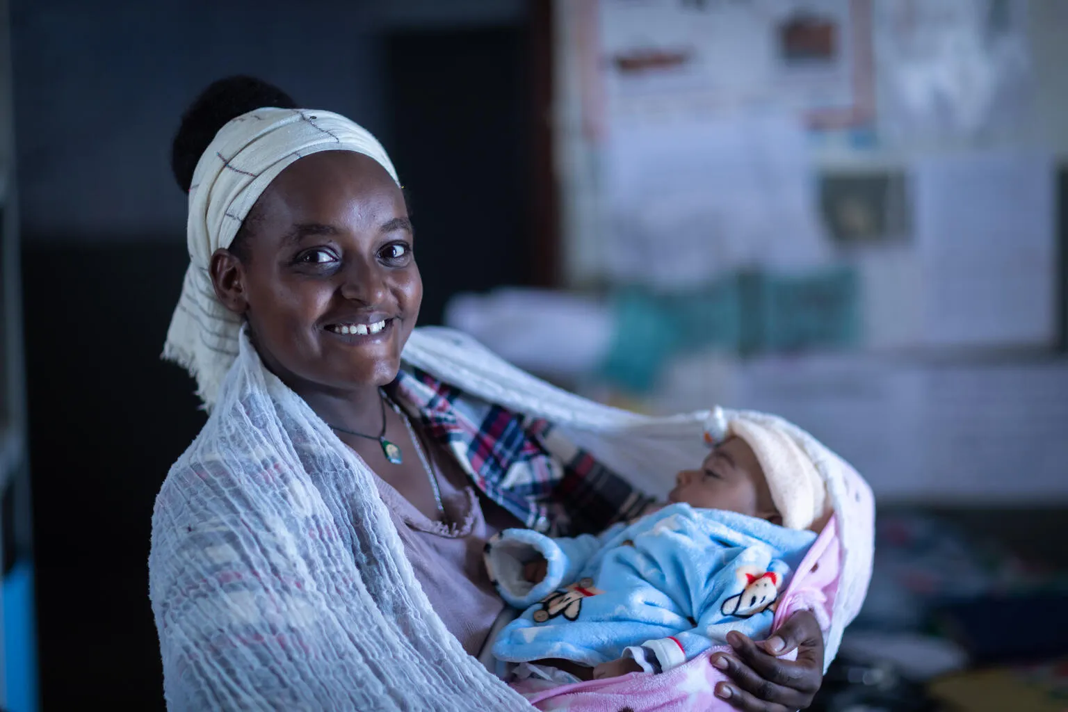 A woman looks at the camera smiling while holding a sleeping baby. 