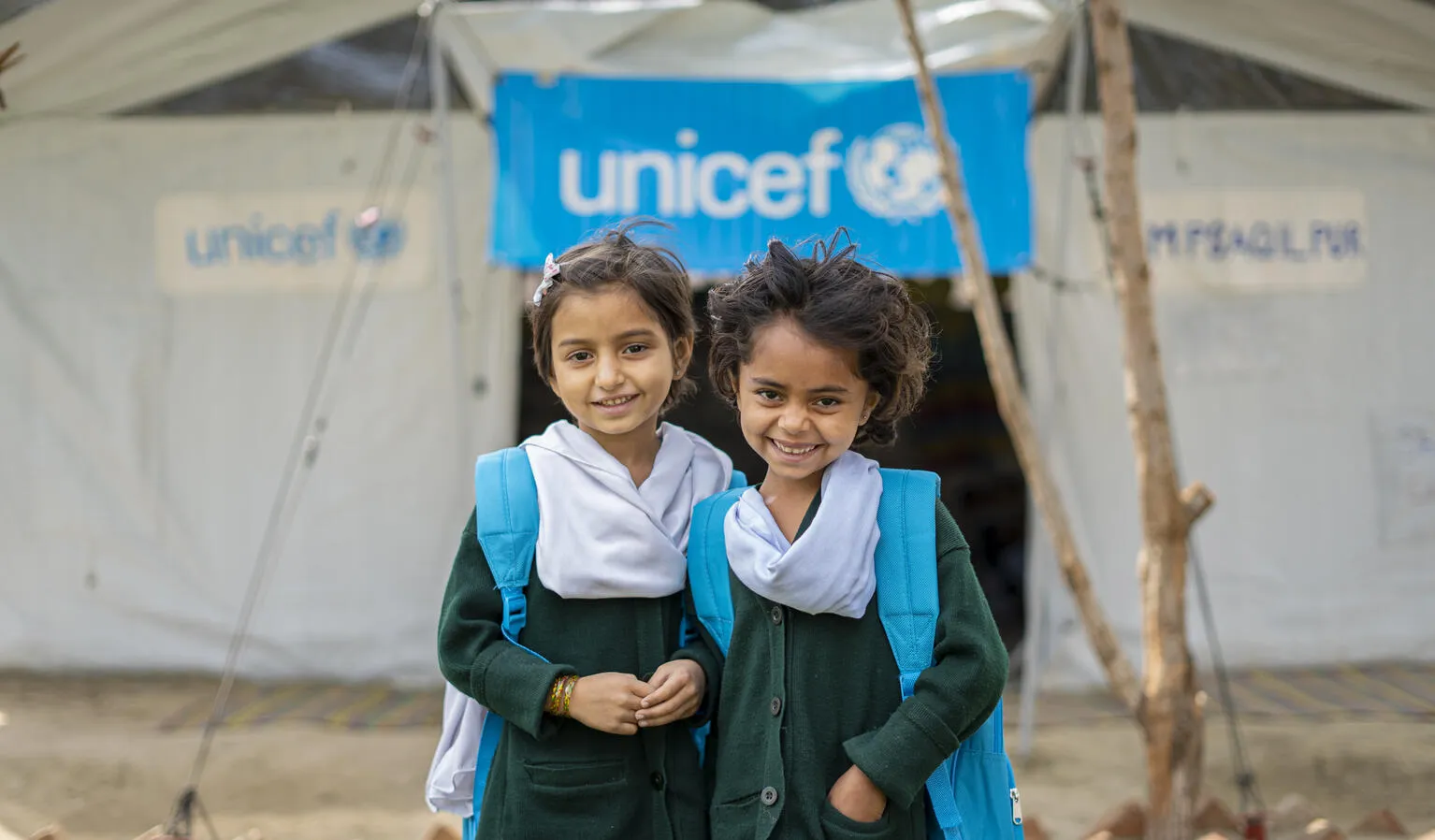 Deux jeunes filles portant des sacs à dos bleus de l’UNICEF devant un centre d’apprentissage temporaire financé par l’organisme sourient à l’objectif.