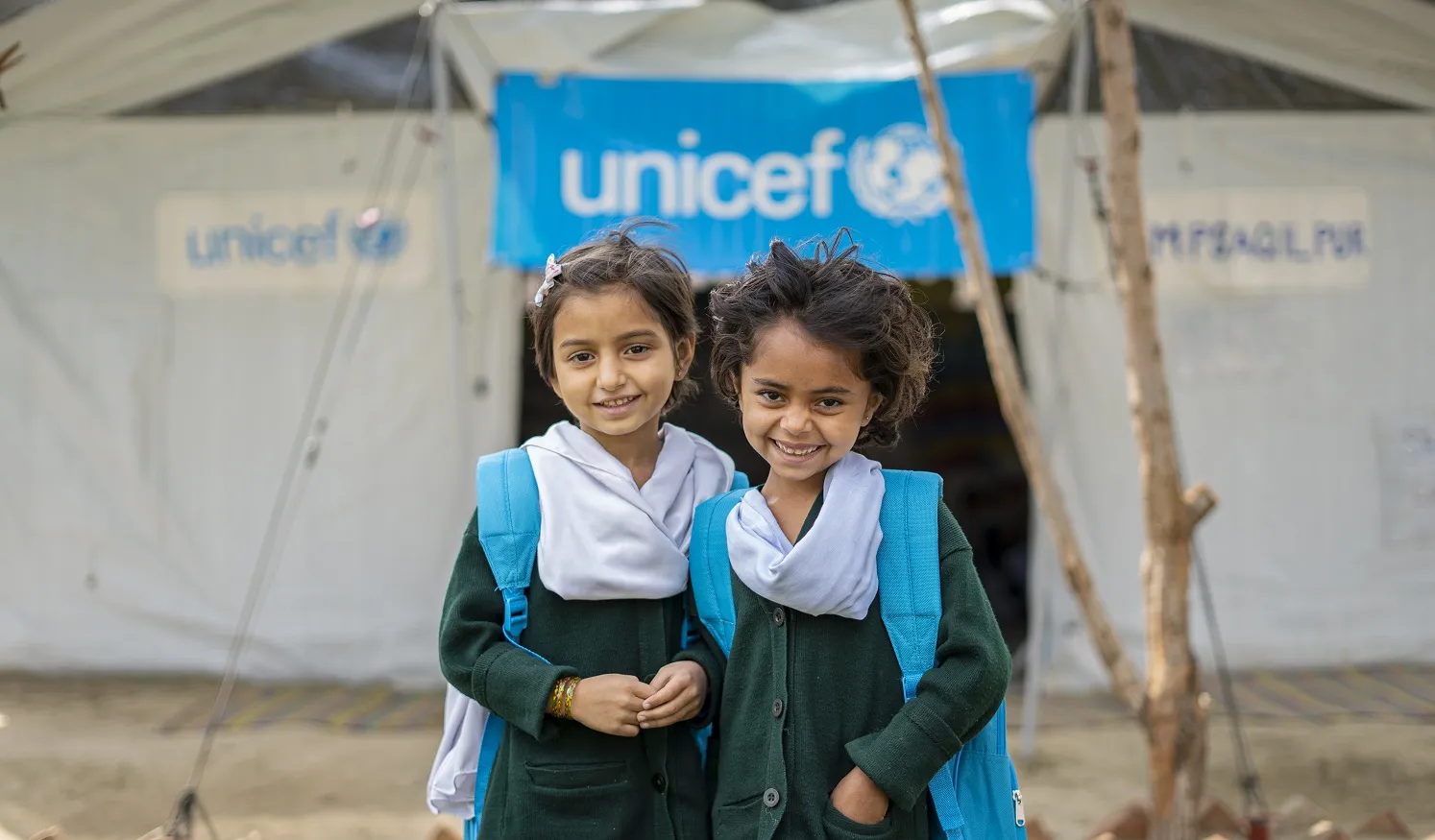Deux jeunes filles portant chacune un sac à dos bleu de l’UNICEF sourient à l’objectif tandis qu’elles se tiennent devant un centre d’apprentissage temporaire financé par l’UNICEF.