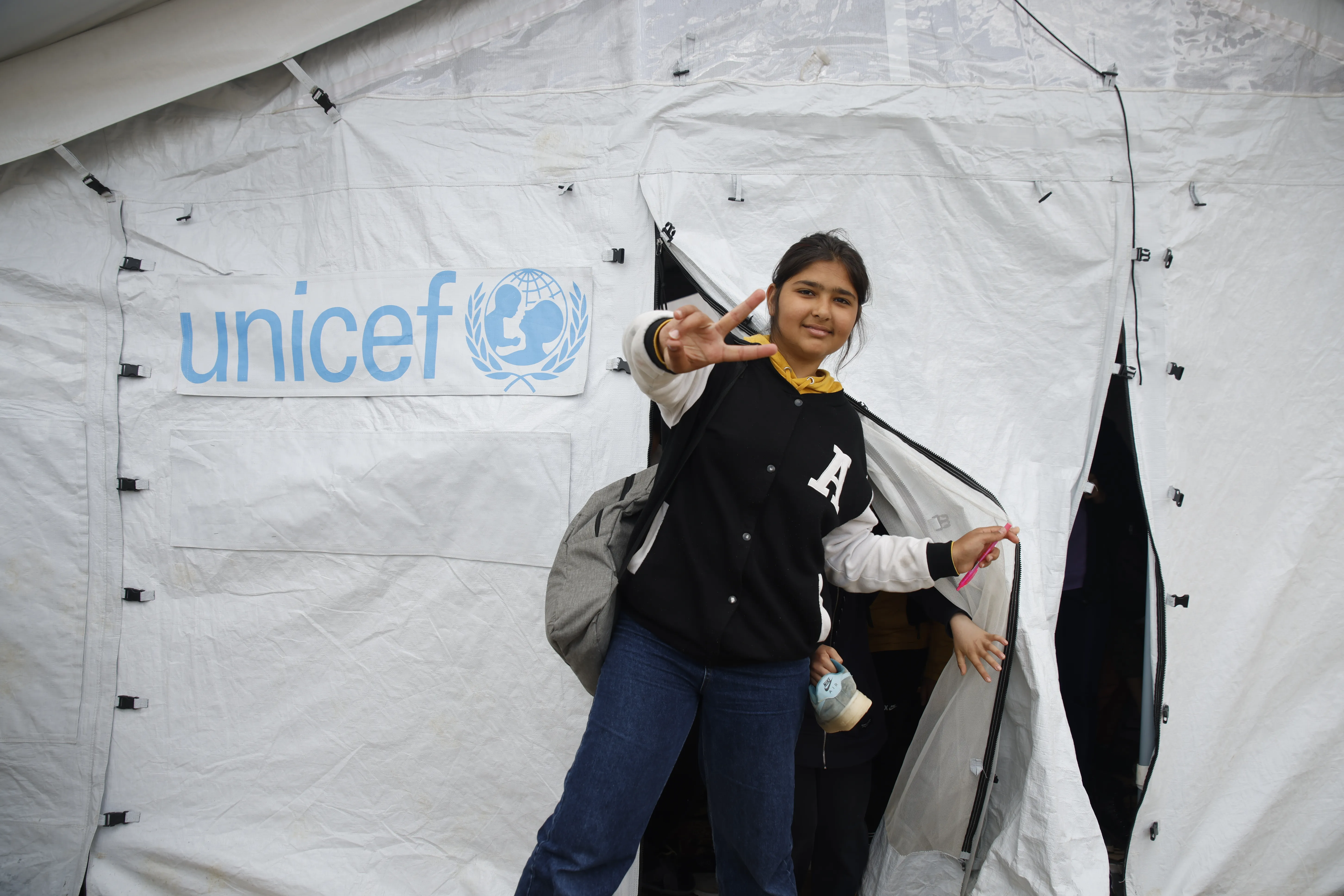 A girl exits a UNICEF tent making the peace gesture with her hand and a bag pack slung over her shoulder.
