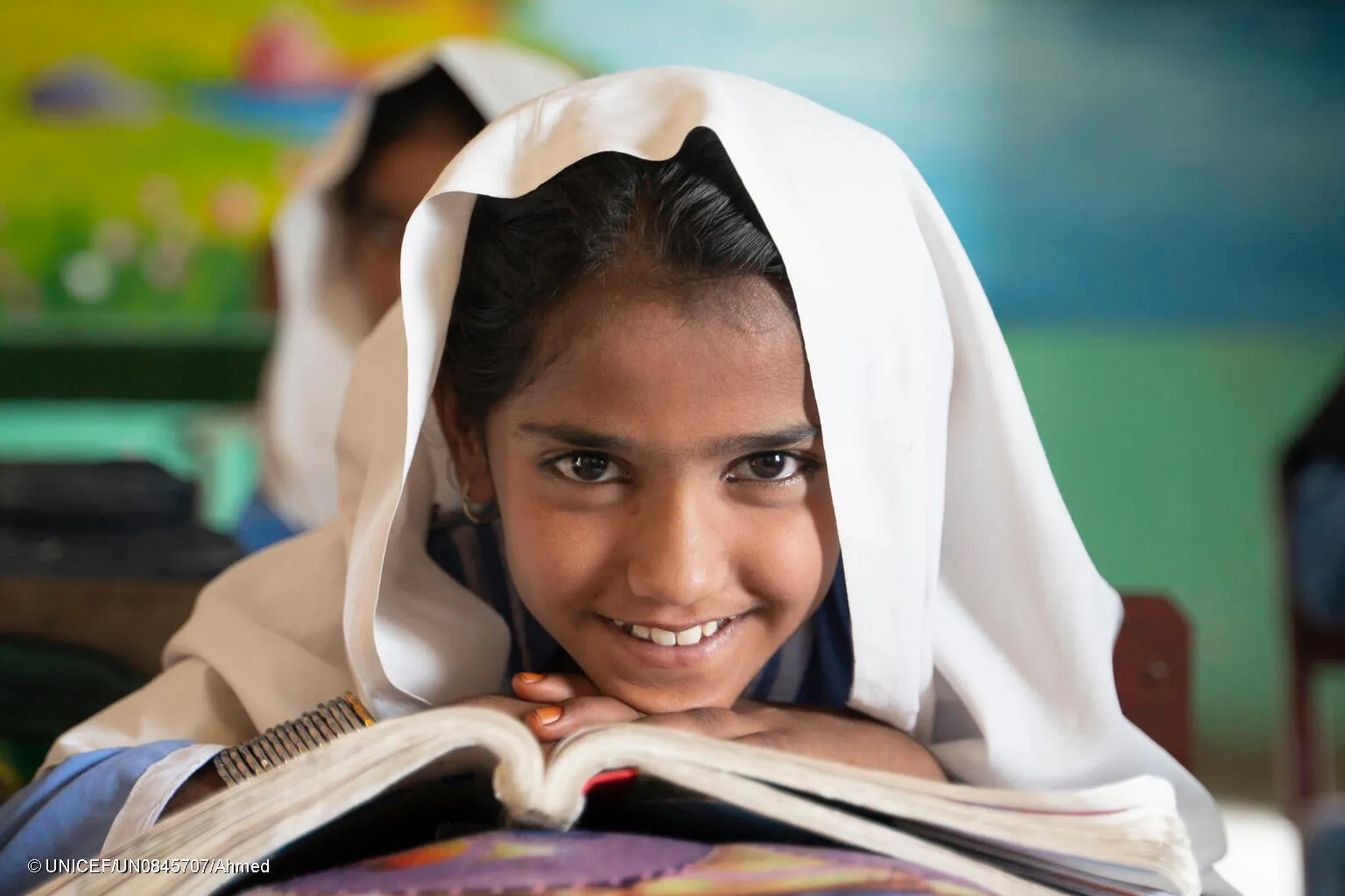 In a classroom in Pakistan, a young girl sitting at her desk smiles with an open book in front of her.  