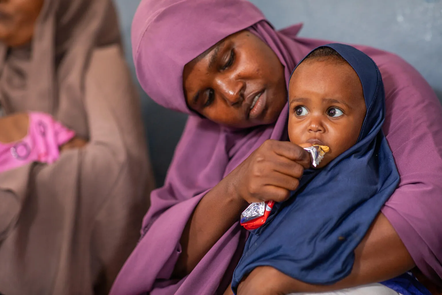 A caring mother in a purple headscarf feeds a young child in a navy hijab, who eats from a snack packet in her arms.