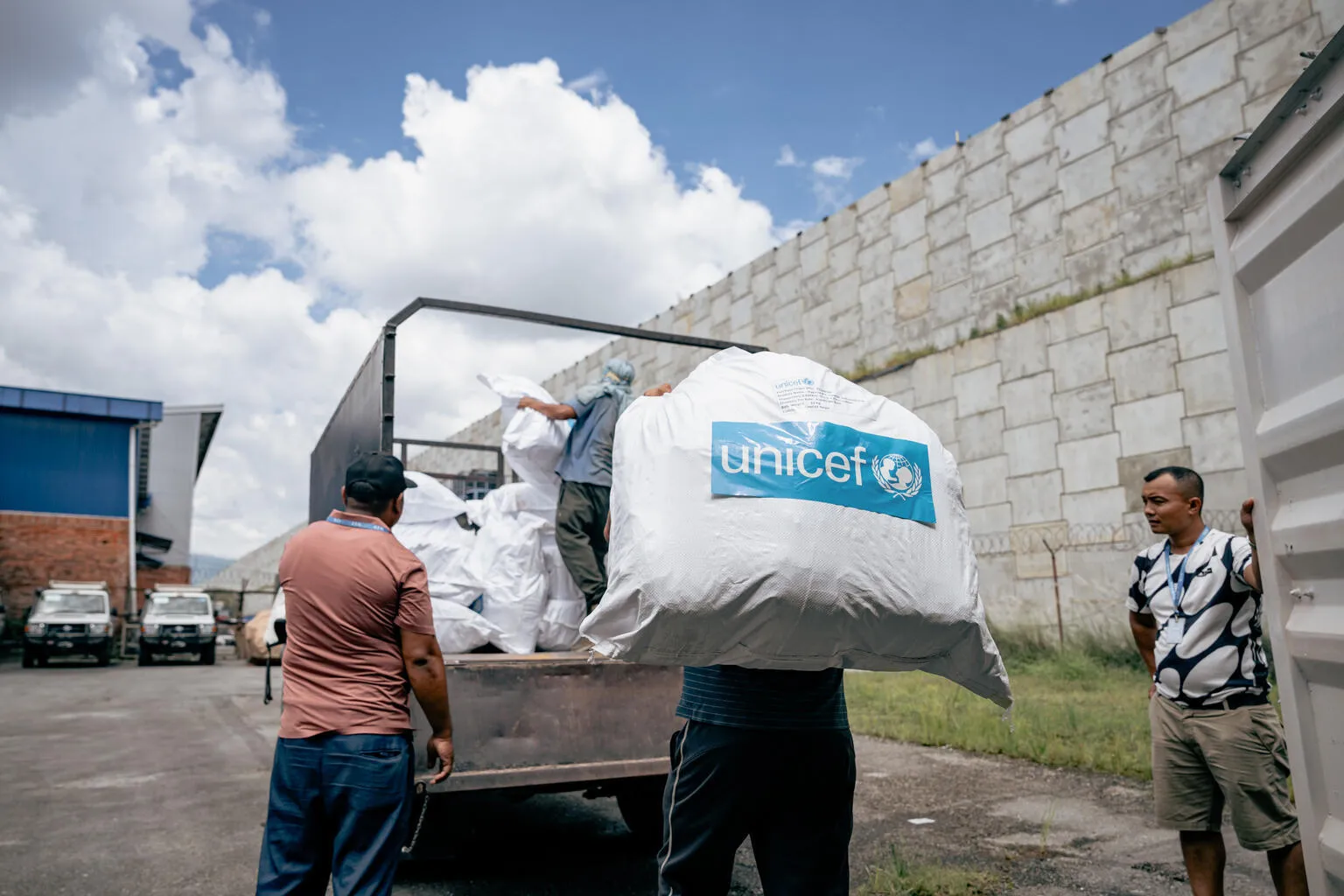 Workers load a large sack with the UNICEF long on it, nto a truck beside a stone wall.