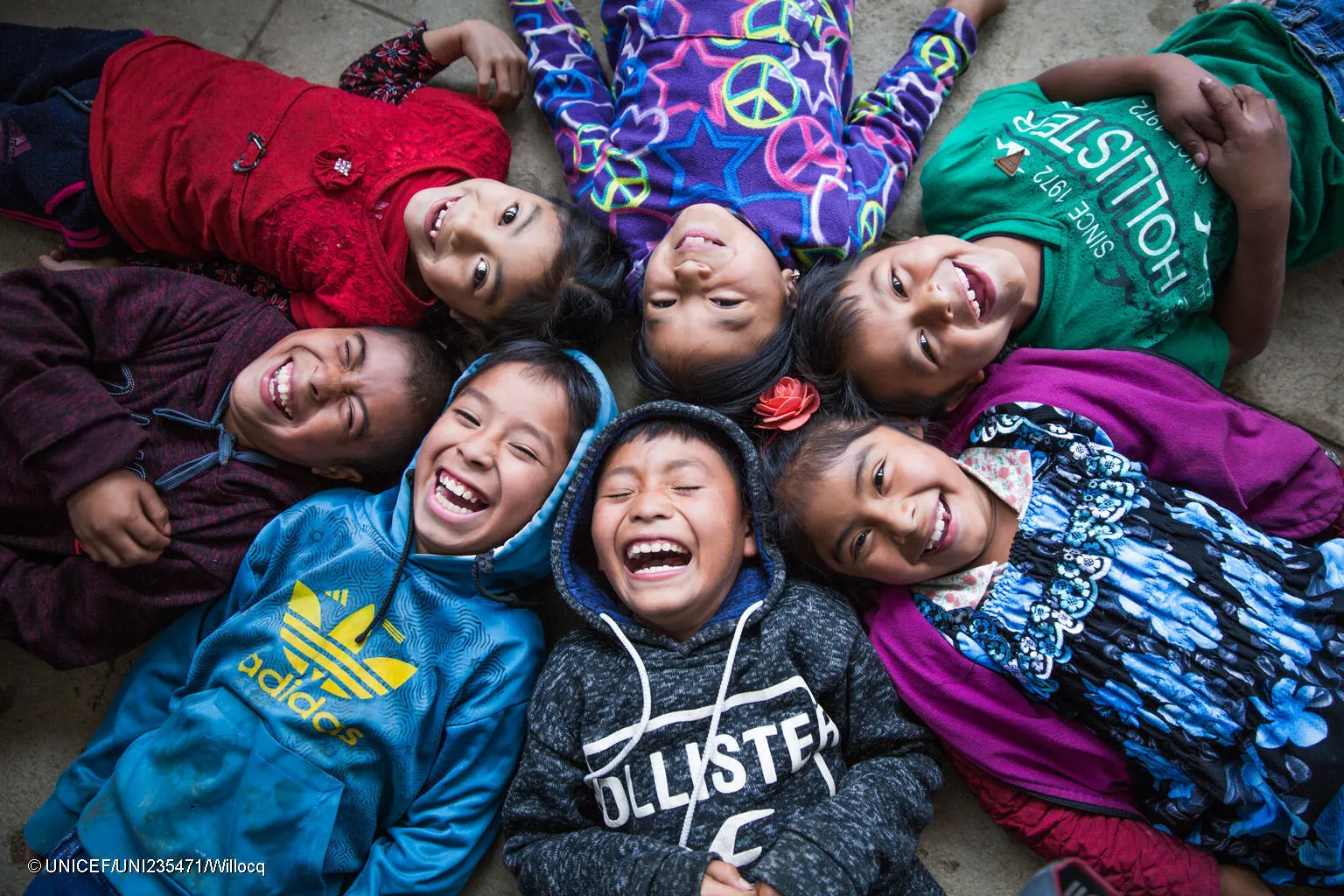 Six children lying on the floor in a circle with their heads touching in. They are looking up at the camera and laughing. 