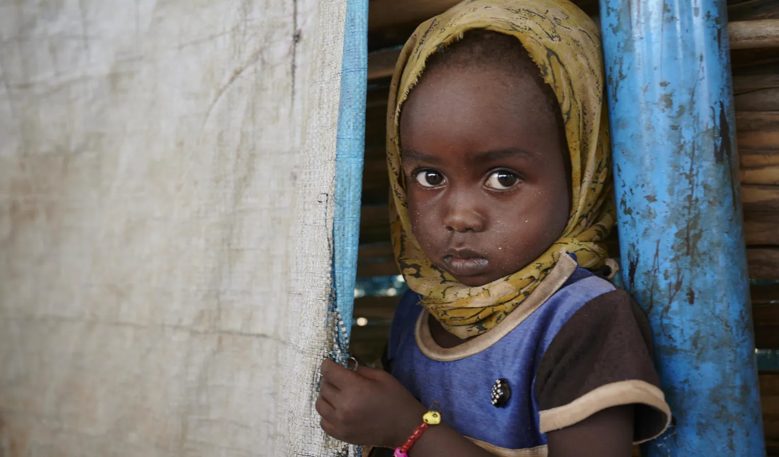 A young child with a serious expression, wearing a yellow headscarf and blue shirt, peeks from behind a curtain