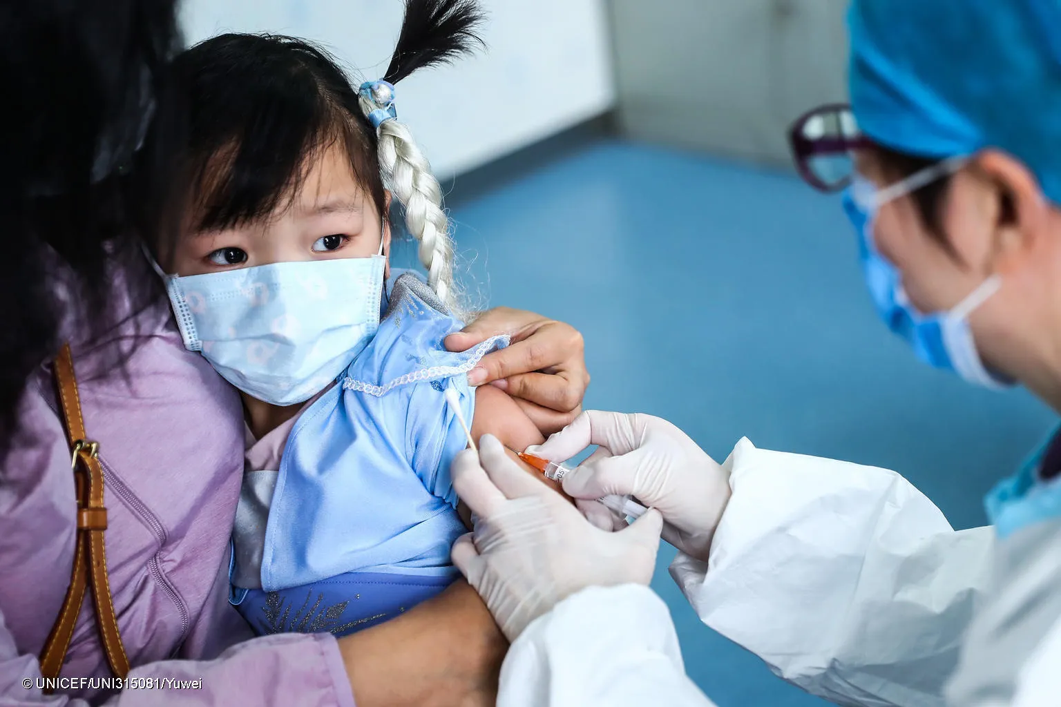 A girl sits in her mother's lap while a health worker administers a vaccine. Both the child and the doctor are wearing masks, the mother's face is not visible. It is a clinic setting. This photo was taken in China in 2020. 