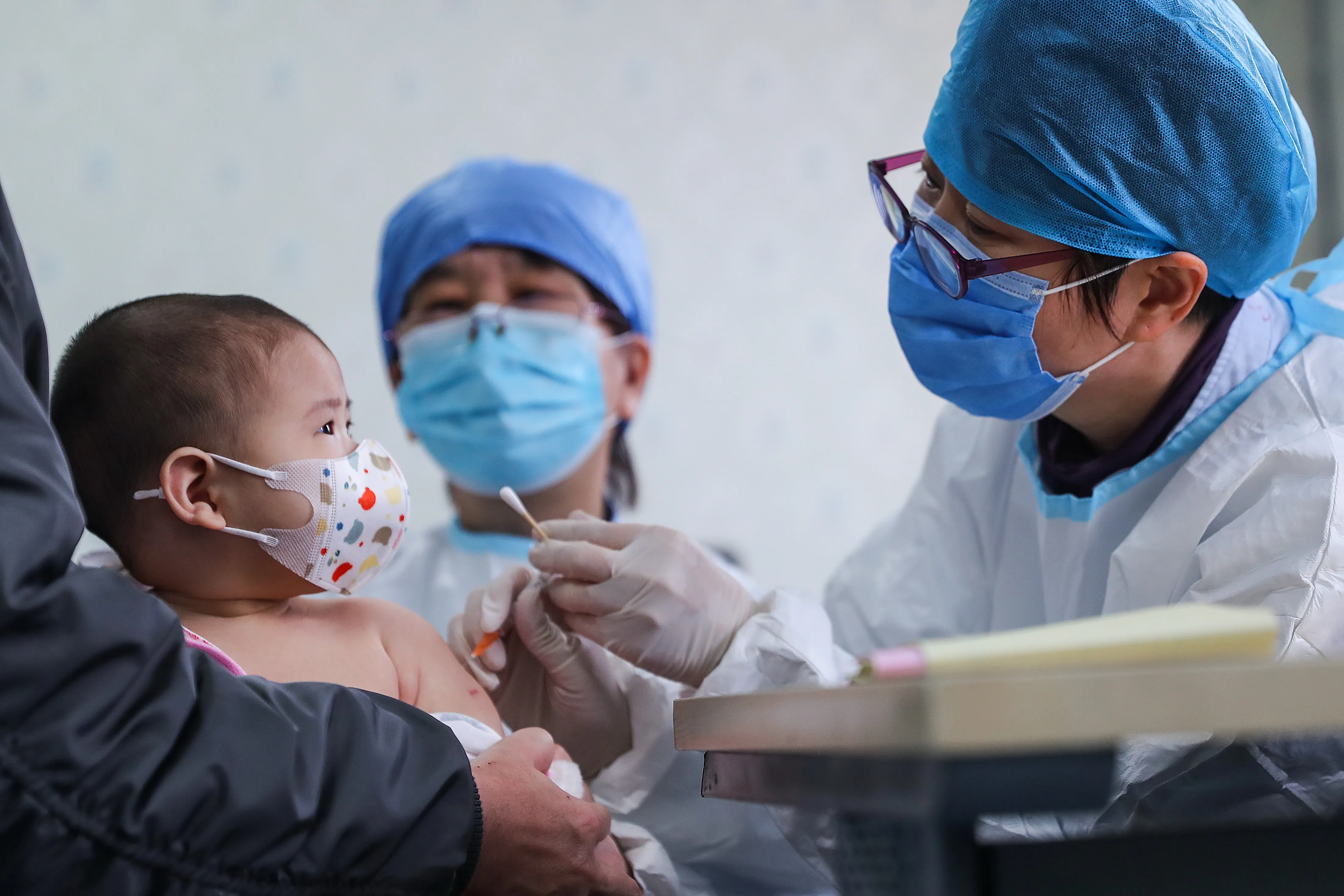 Baby wearing mask looking a doctor in a cap and mask holding a cotton swab with gloved hands