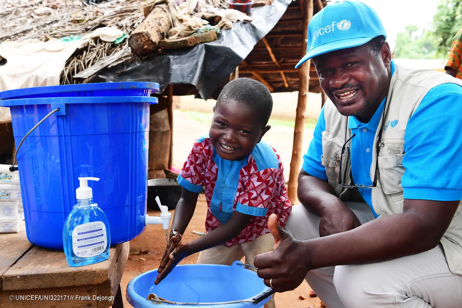 A man and a child squat in front of a bucket with a tap, the boy is washing his hands and the man is giving a thumbs up sign. The man is wearing a UNICEF cap and vest. 